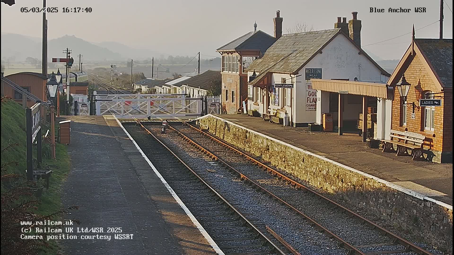 A sunny view of a train station with two train tracks and low stone walls, including a signal box and passenger shelters.