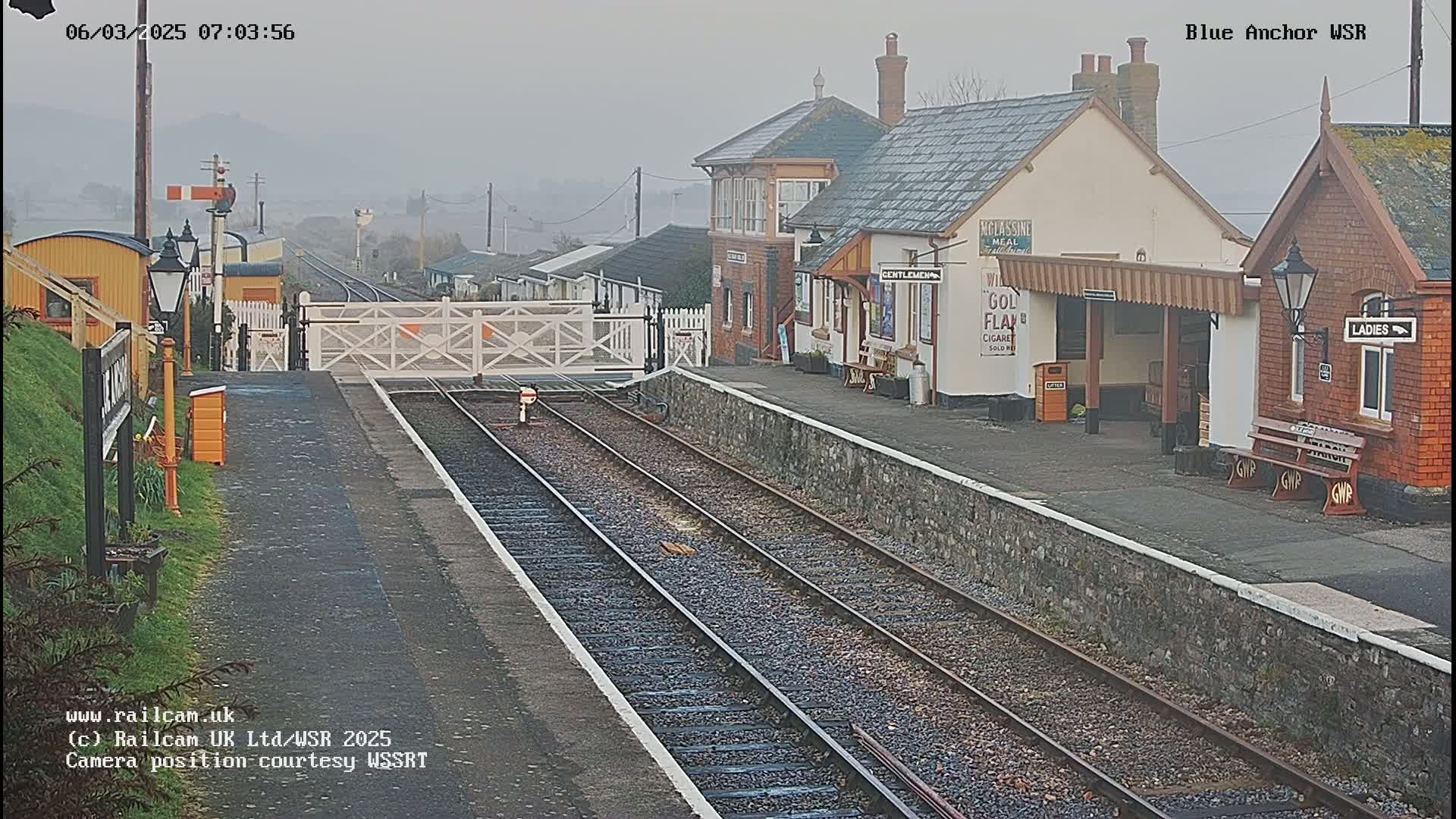A foggy view of a train station platform with two sets of tracks and several buildings, including a signal box and waiting shelters.