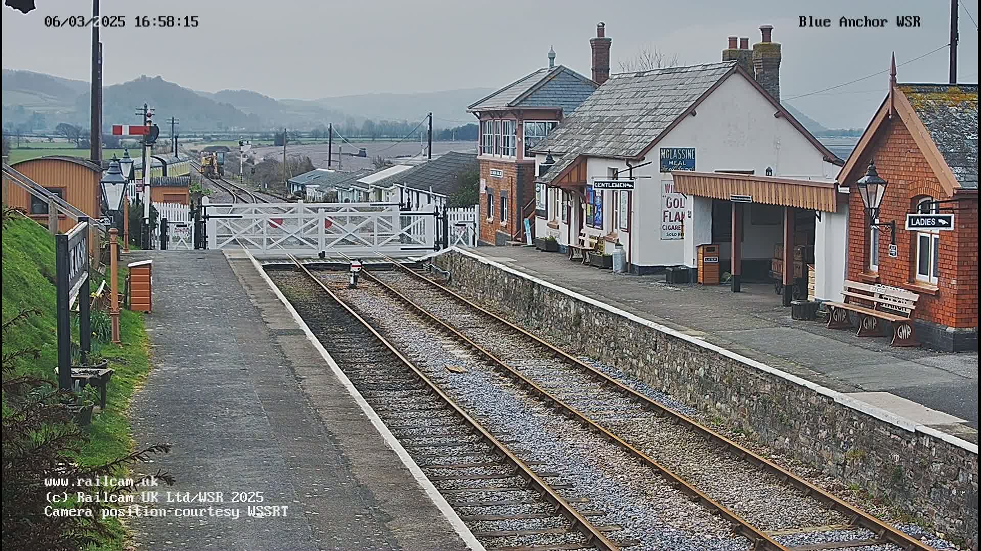 A train station with several buildings, a closed railway crossing gate, and train tracks under an overcast sky.