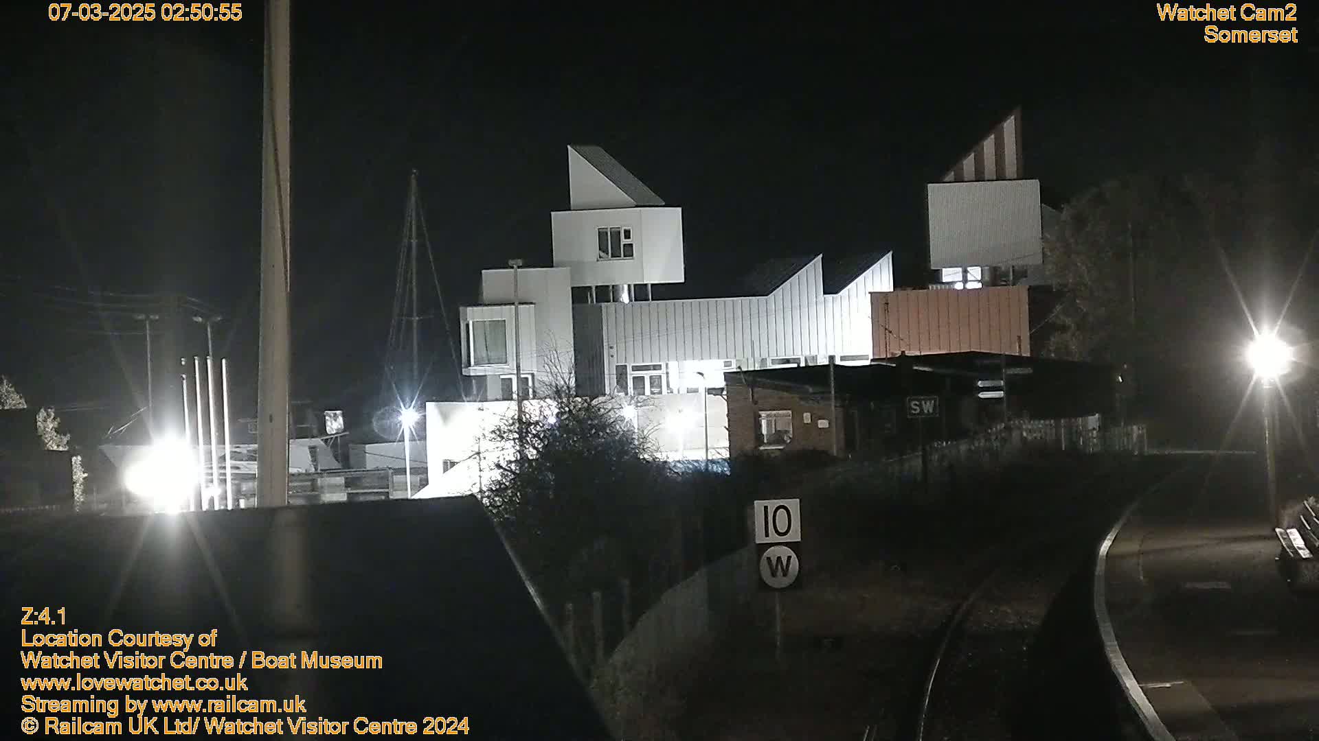 A modern, multi-level white building is illuminated at night, next to a railway track with a speed limit sign.