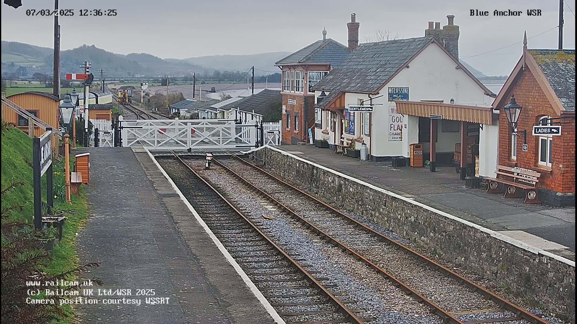 A train station with a low brick building and a covered platform is viewed from the tracks on an overcast day.