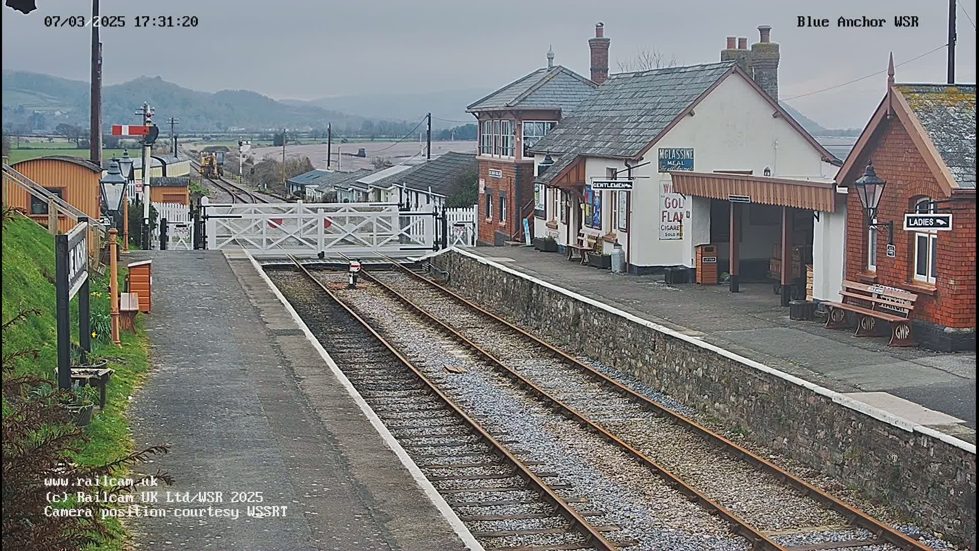 A train station with two platforms and a signal gate, under an overcast sky, is situated beside buildings, with hills visible in the background.