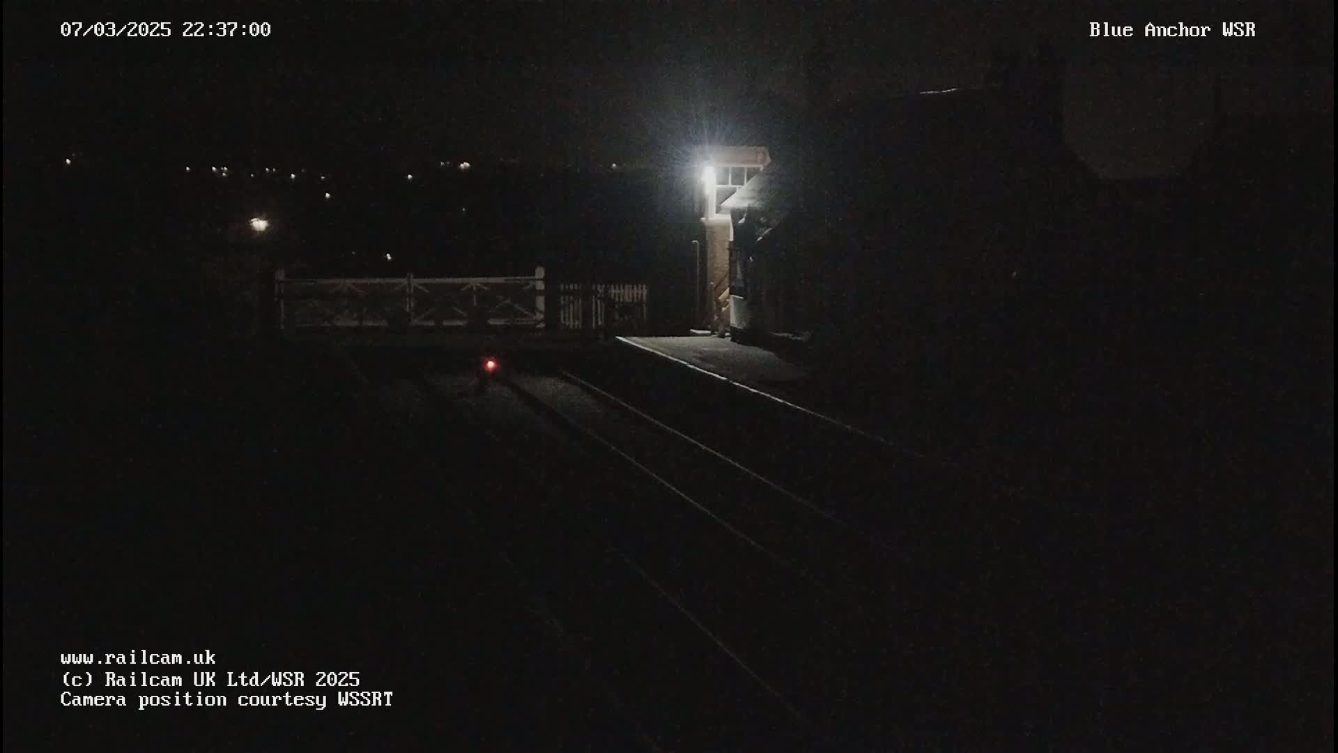A dimly lit nighttime train platform with train tracks and a small building is visible under a dark sky.
