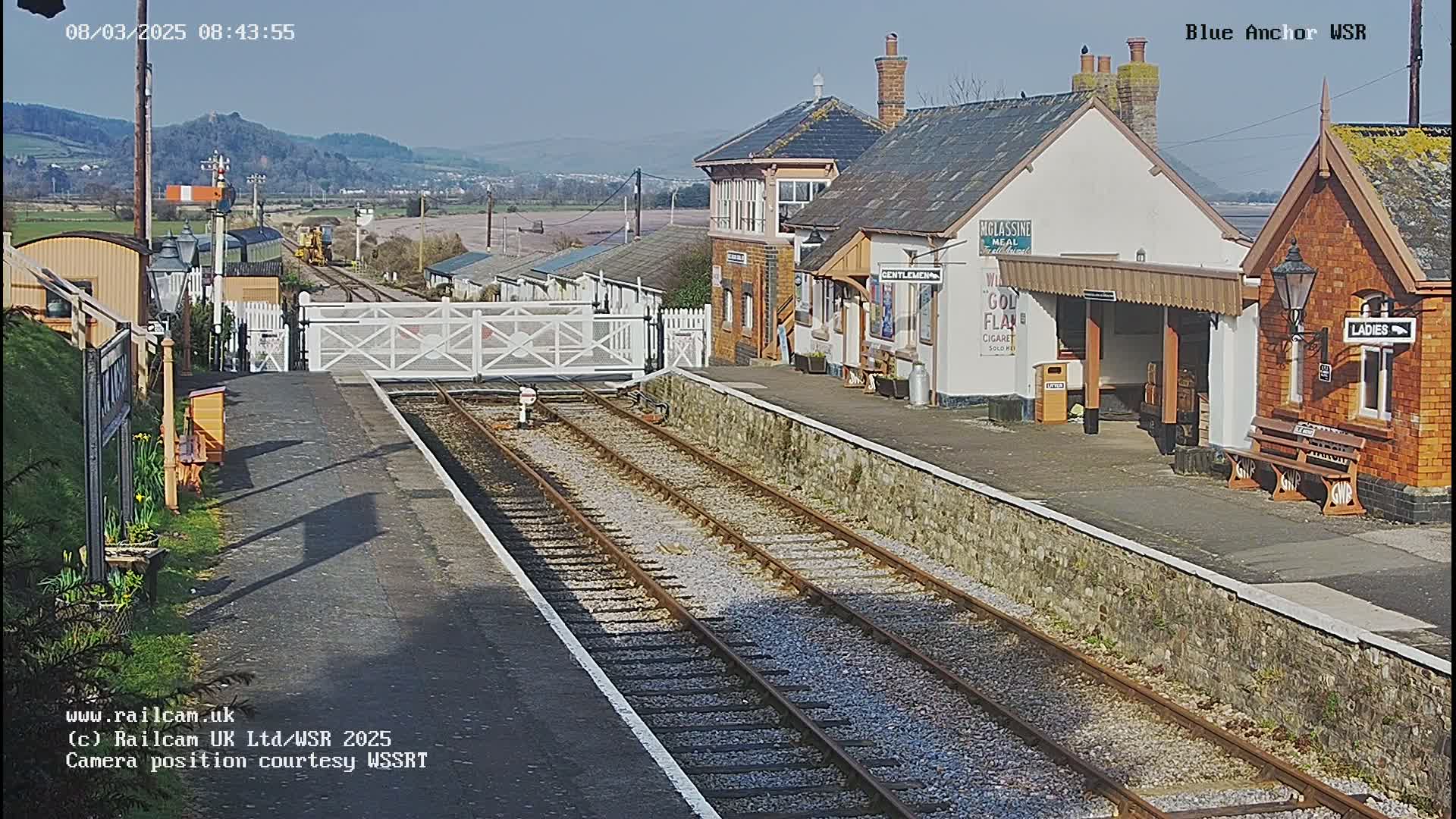 A sunny day at a train station with two sets of tracks, several buildings, and a level crossing gate.
