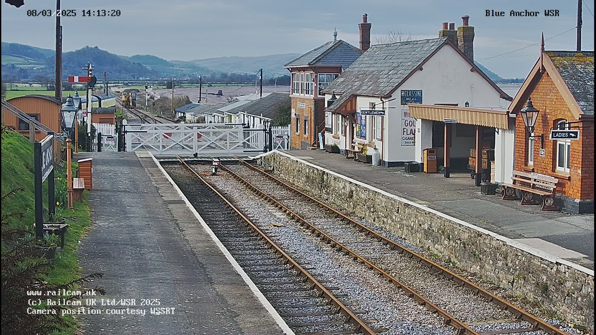 A railway station with train tracks, a signal box, and several buildings is viewed on an overcast day.