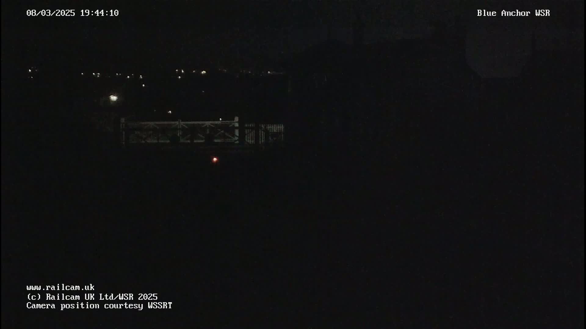 A dark nighttime scene shows a dimly lit bridge and distant lights under a clear, black sky.