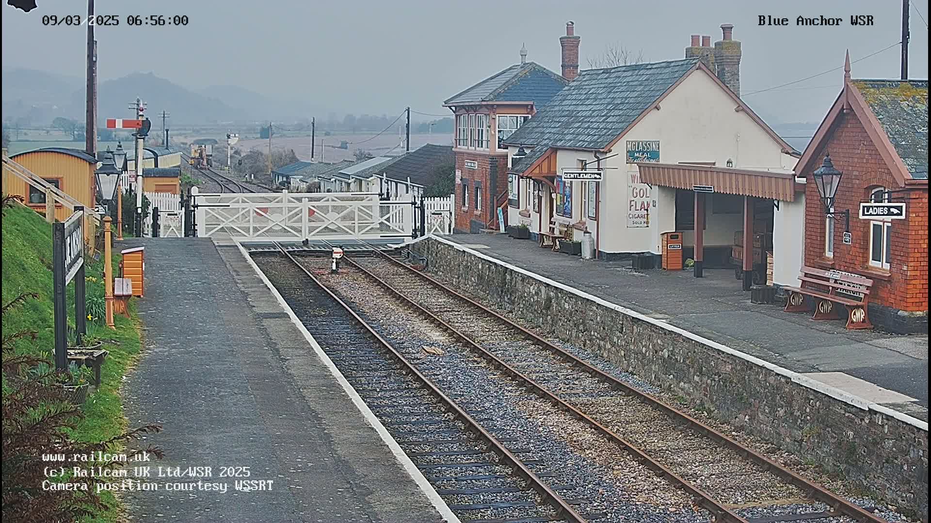 A train station with a brick building and covered platform, under an overcast sky, has two sets of train tracks extending from it.