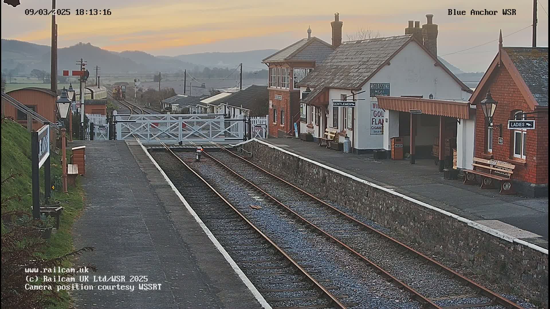 A train station with a signal box and several buildings, under a soft, dusky sky, with train tracks extending into the distance.