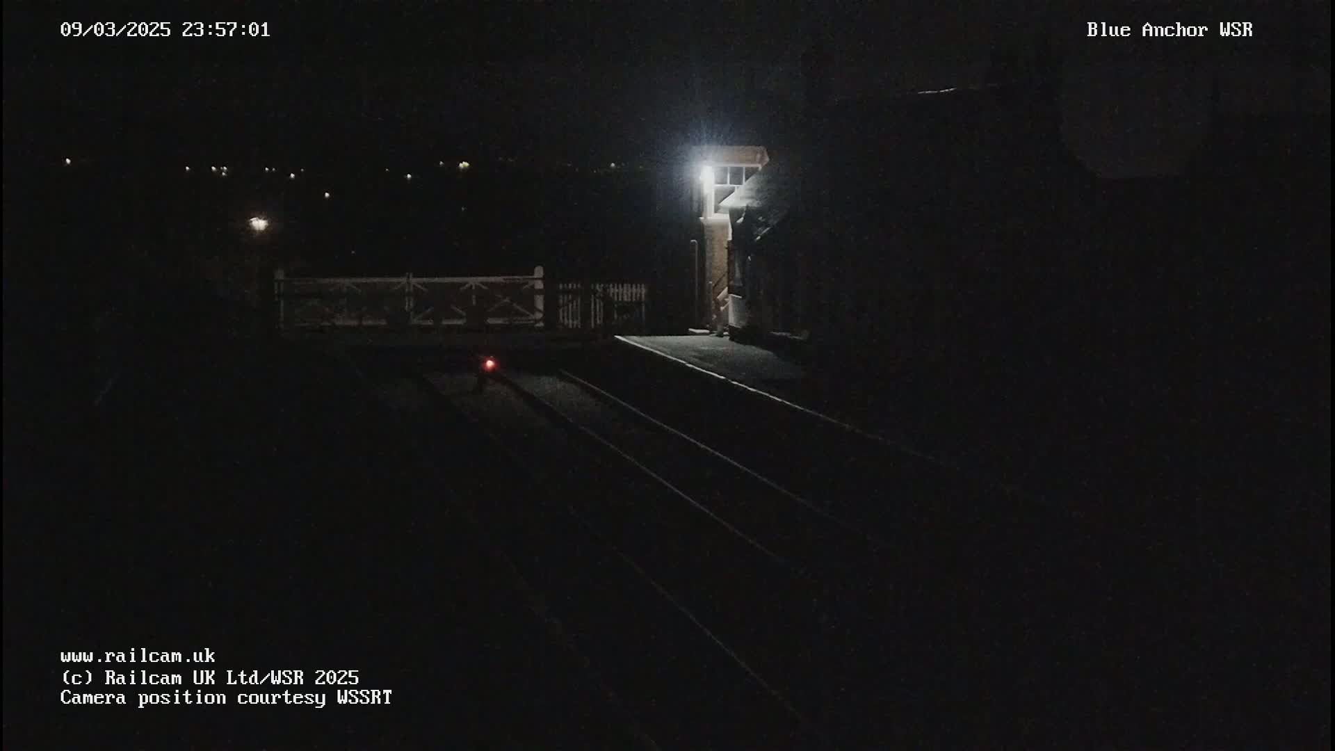 A dark nighttime view of a train station platform with train tracks, a building, and a single light illuminating the scene.