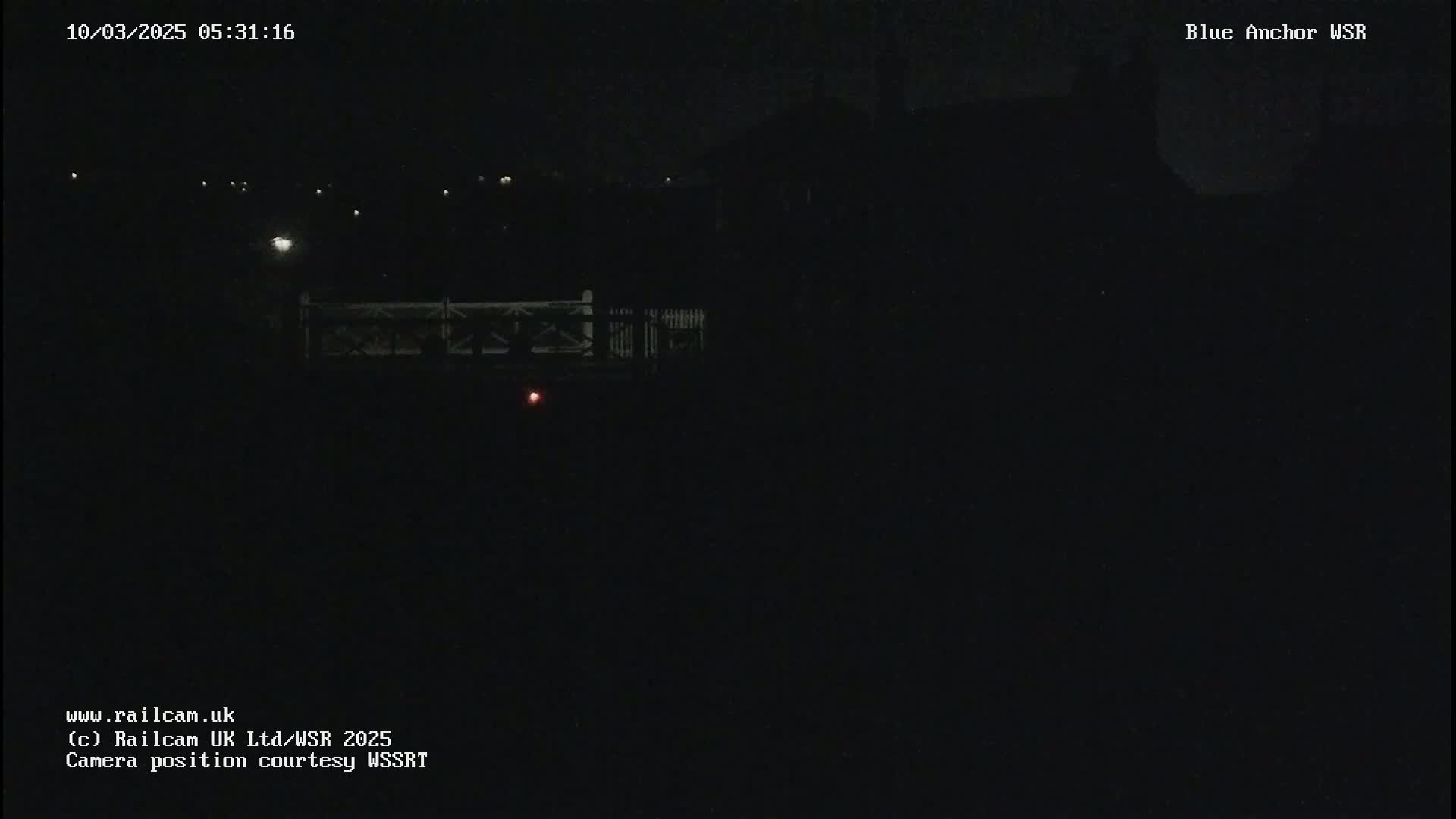 A nighttime view shows a dark landscape with a dimly lit bridge and a faint red light in the distance.