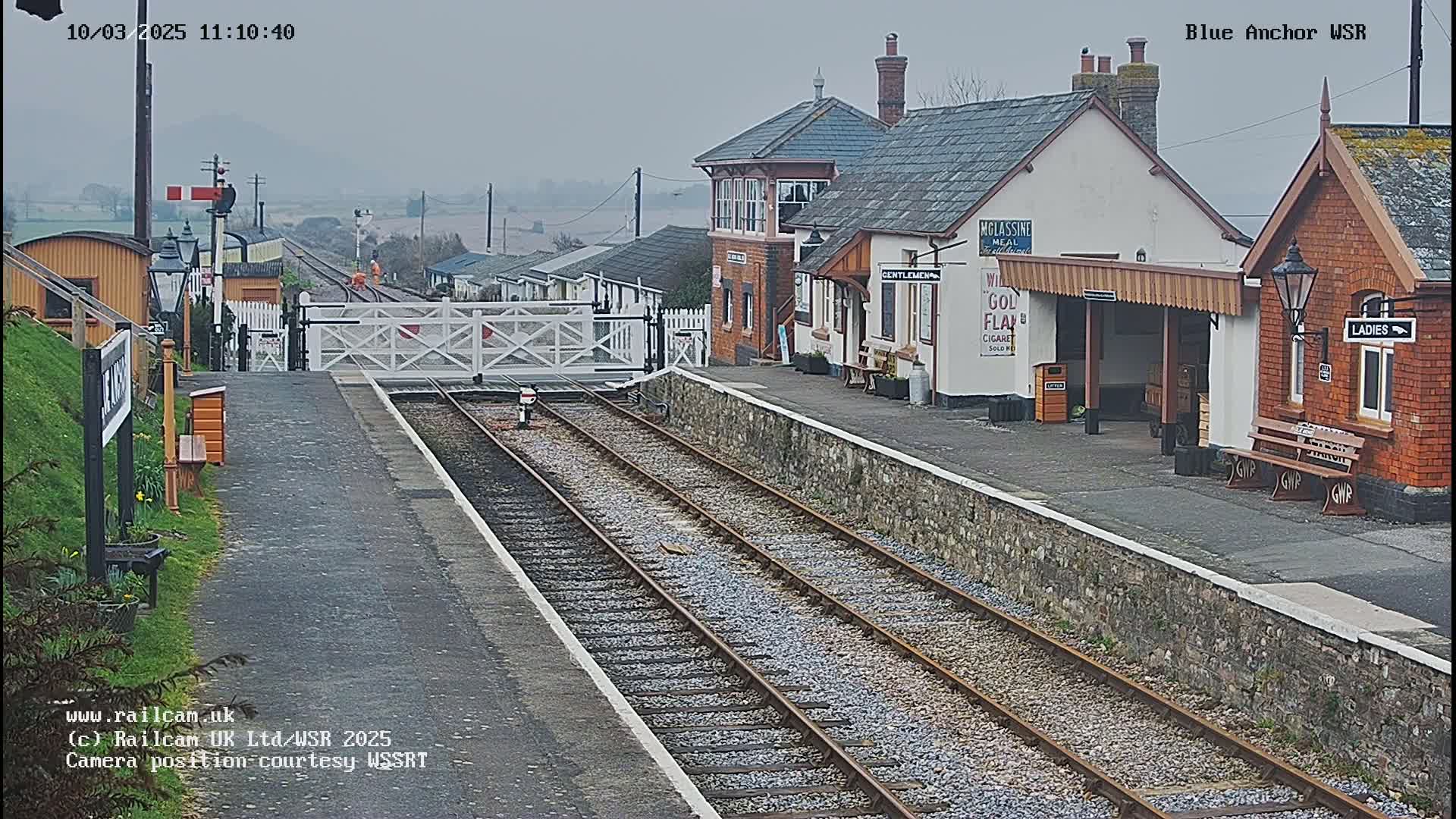 A train station platform with a railway track, station buildings, and level crossing gates under an overcast sky.