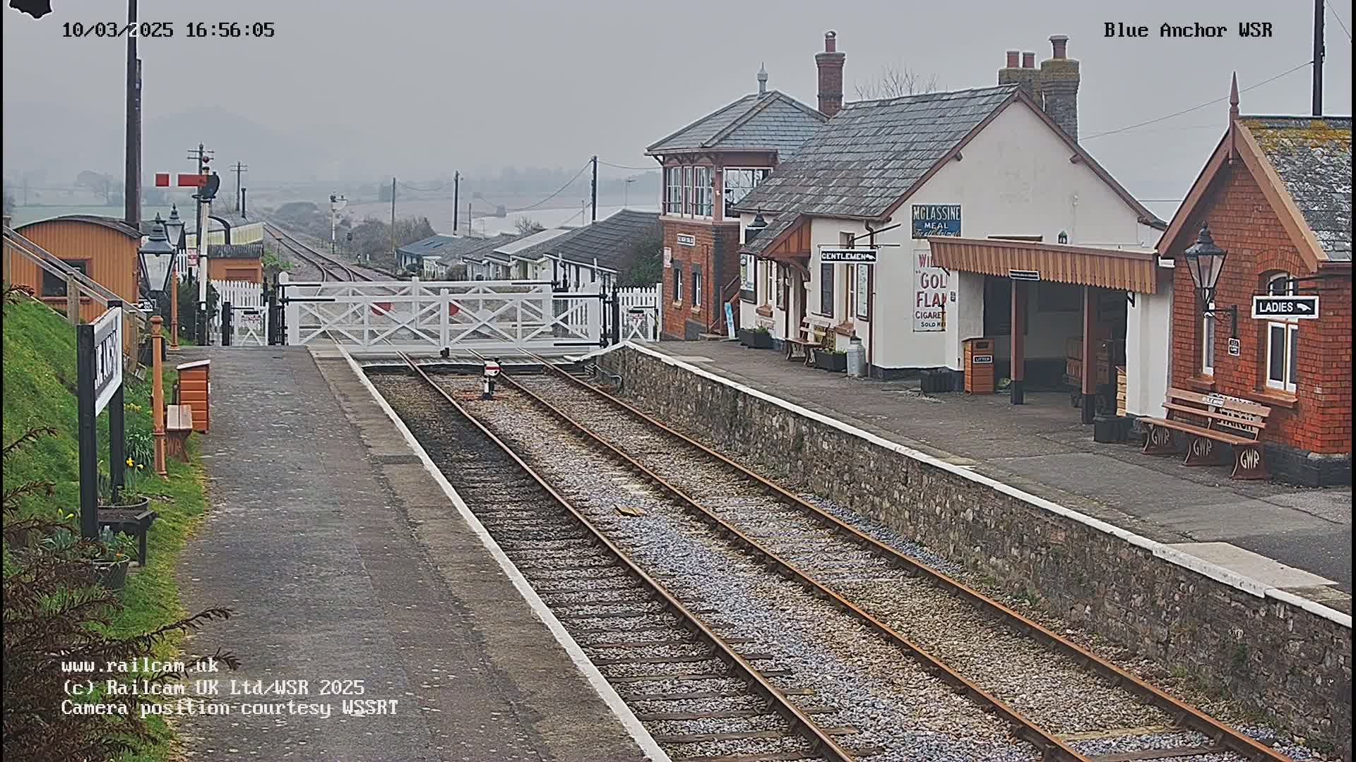 A train station with two tracks and several buildings is visible on an overcast day.