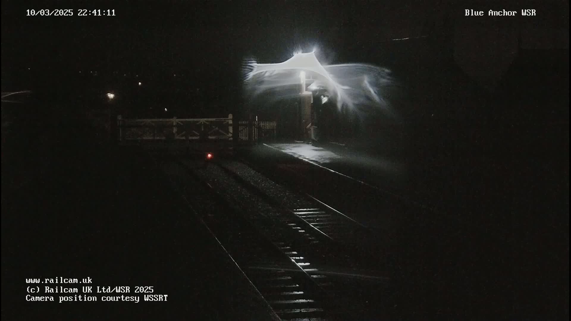 A nighttime view of a train platform with snow blowing intensely around a lit lamppost, illuminating the tracks and platform.