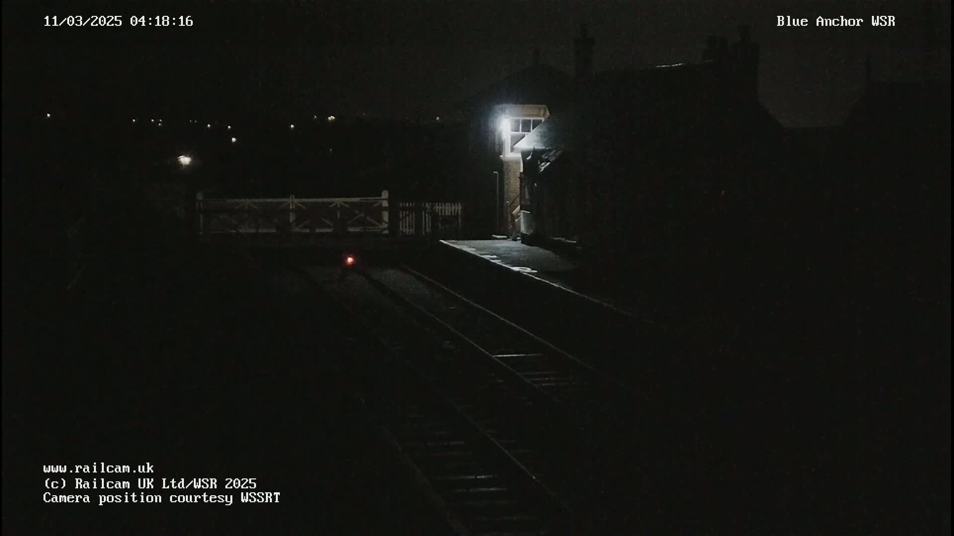 A dimly lit train station platform at night is shown with train tracks extending into the darkness.