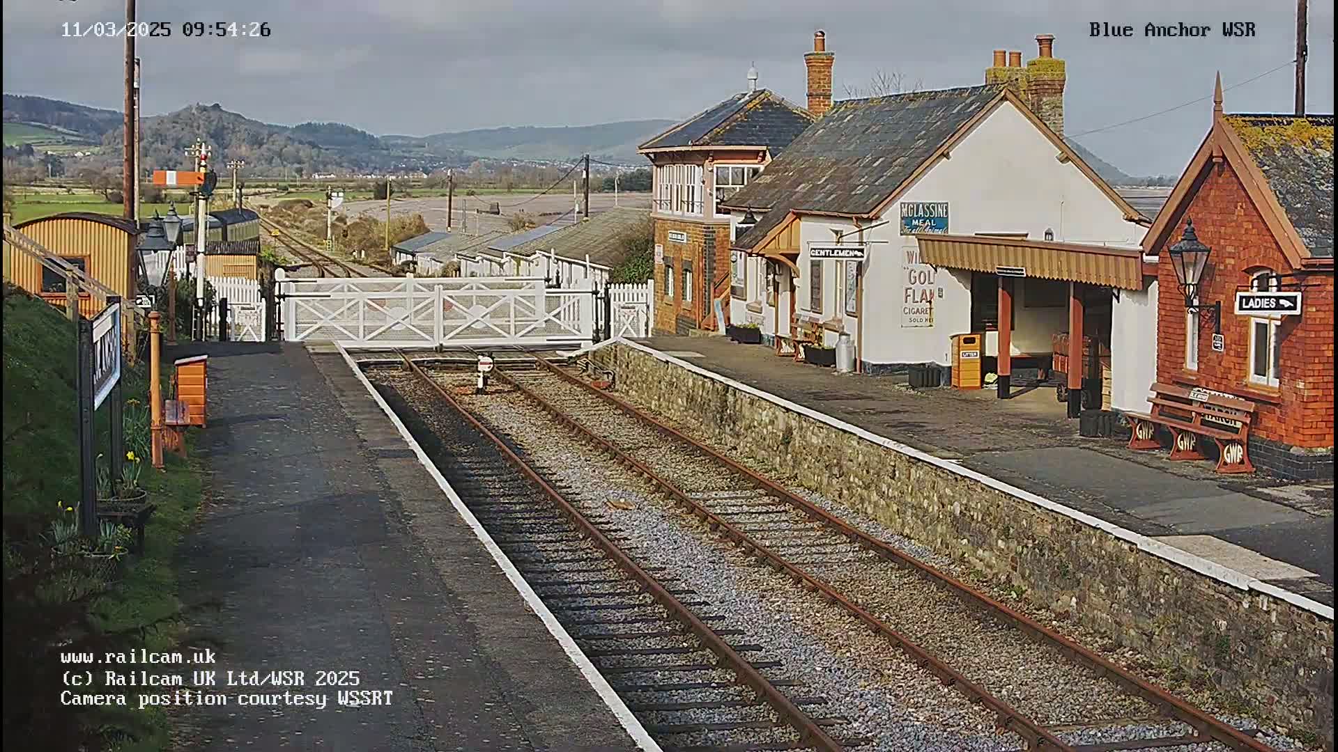 A train station with a few buildings, train tracks, and a level crossing, all under a partly cloudy sky.