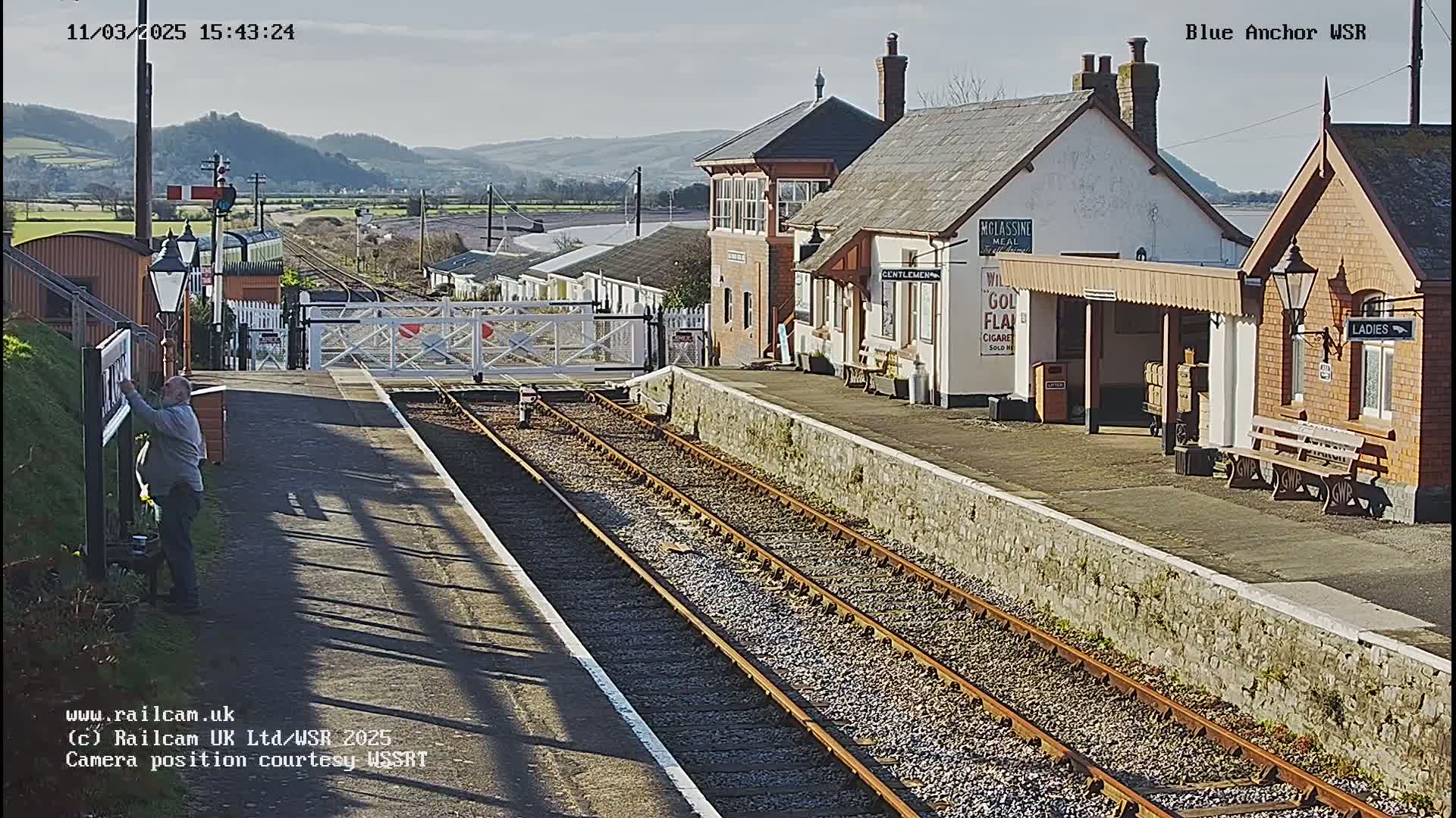 A sunny day at a train station with a low stone wall separating the platform from the tracks, buildings, and a person working near a sign.
