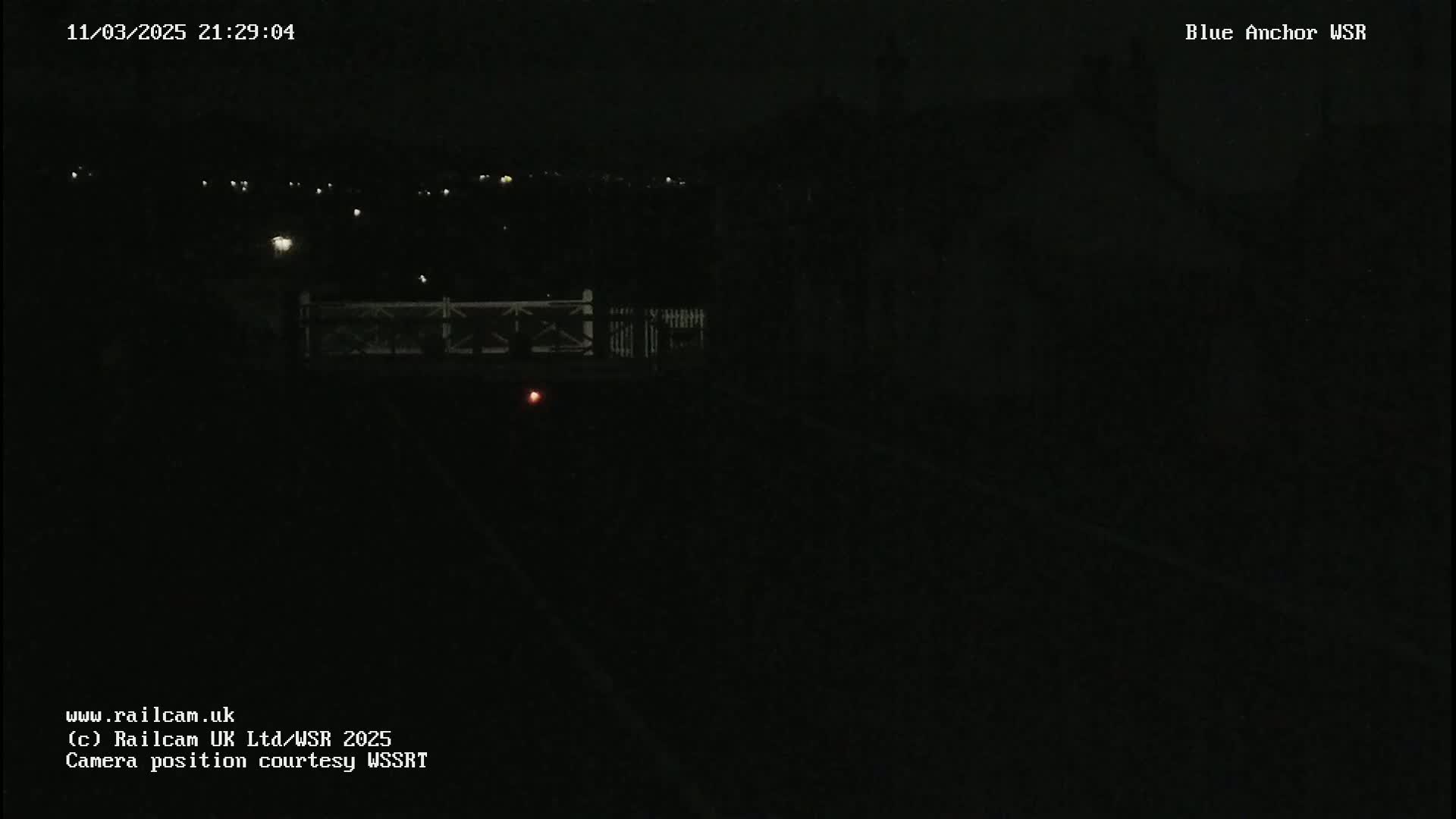 A dark nighttime scene shows a dimly lit area with a fence and a faint red light in the distance.
