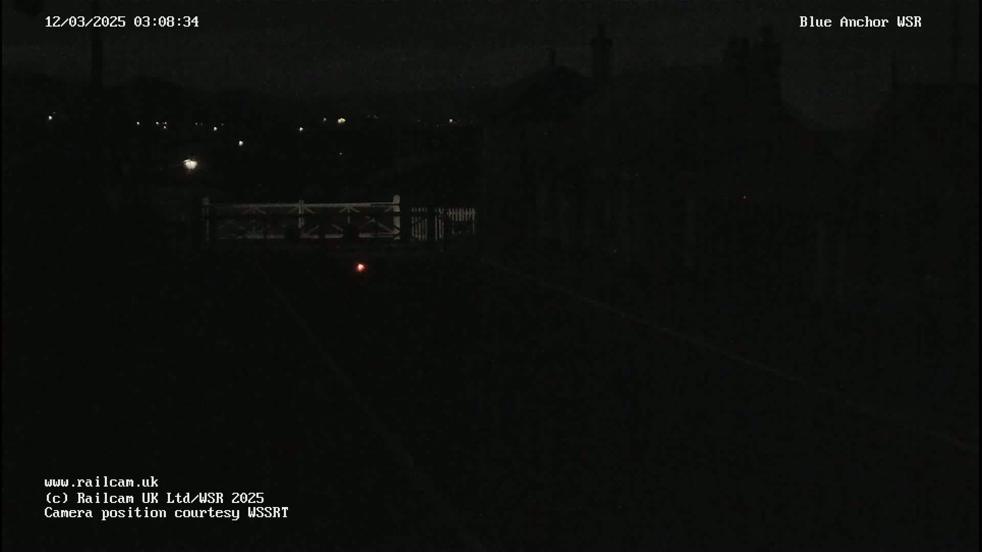 A dark, nighttime scene shows a railway crossing gate and a dimly lit road or track leading to a dark building in the distance under a mostly clear, night sky.
