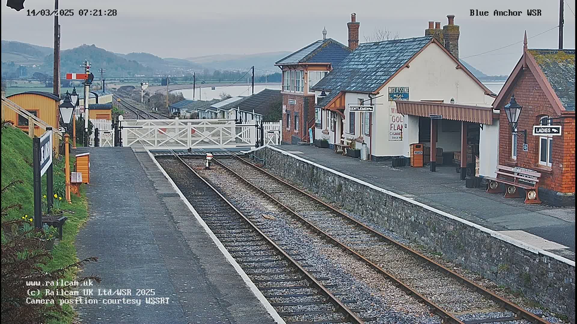 A train station with a signal box and waiting shelters is viewed from the tracks on an overcast day.