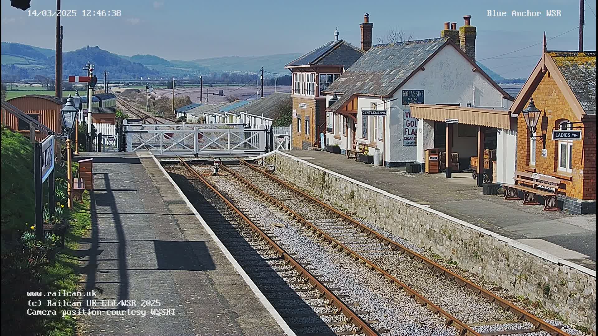 A sunny day at a quaint train station with a signal gate, two sets of train tracks, and several historic-looking buildings.