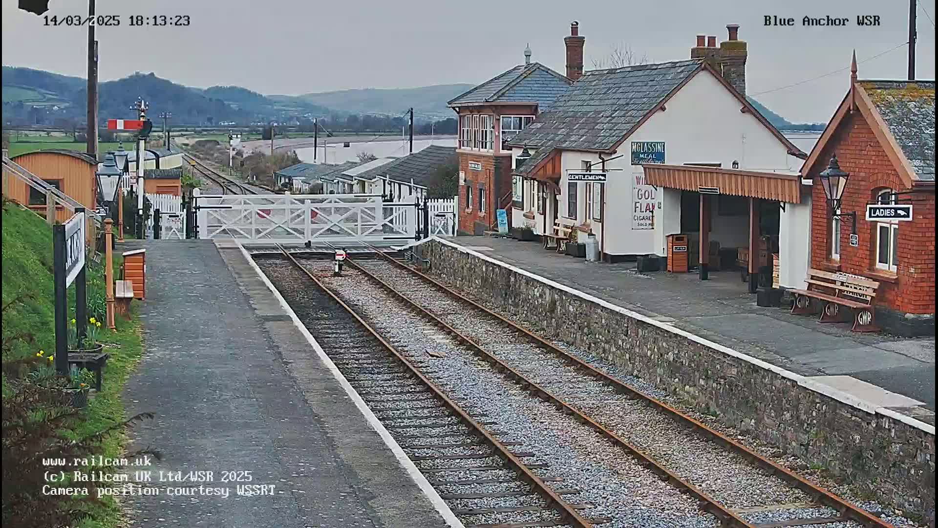A railway station with two platforms and a signal box is visible under an overcast sky; a body of water is in the distance.
