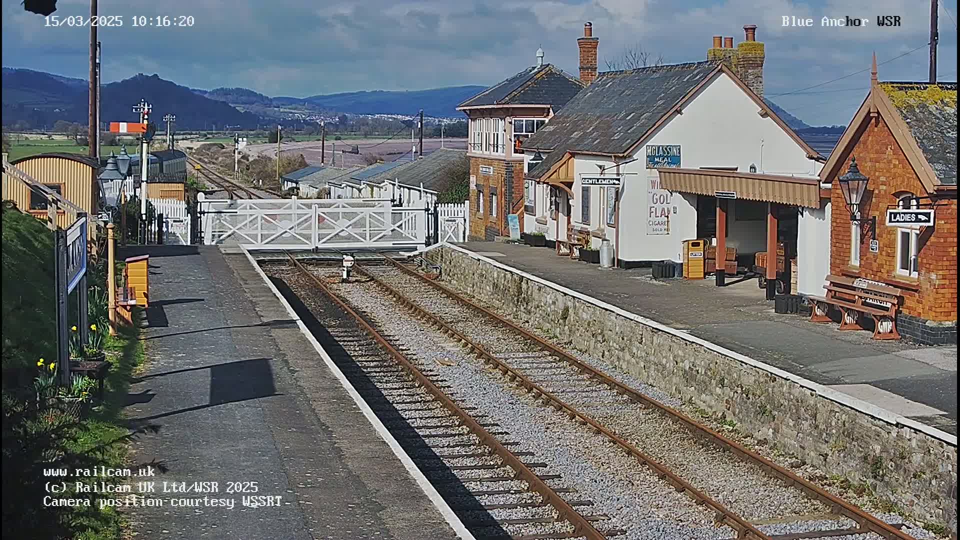 A sunny day at a train station with a brick building, a signal gate, and train tracks extending into the distance, beyond which are hills under a partly cloudy sky.