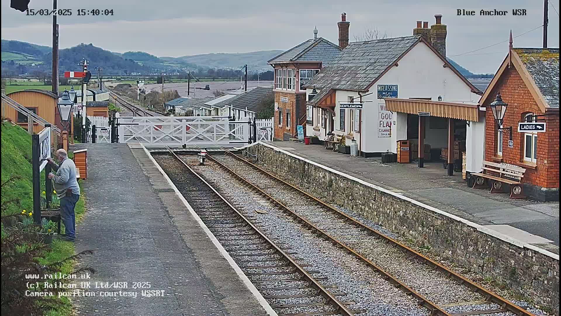 A railway station with a brick building and a signal box, under an overcast sky, shows multiple train tracks and a person working near a sign.