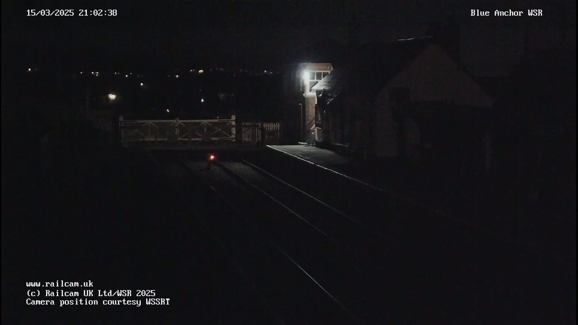 A dark nighttime view of a train station platform with a building and train tracks.