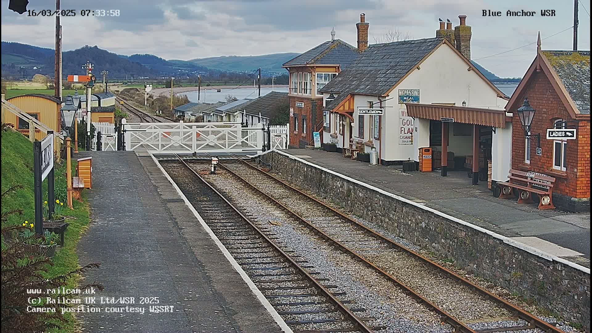 A train station with a brick building and a covered waiting area is viewed from the tracks on an overcast day.