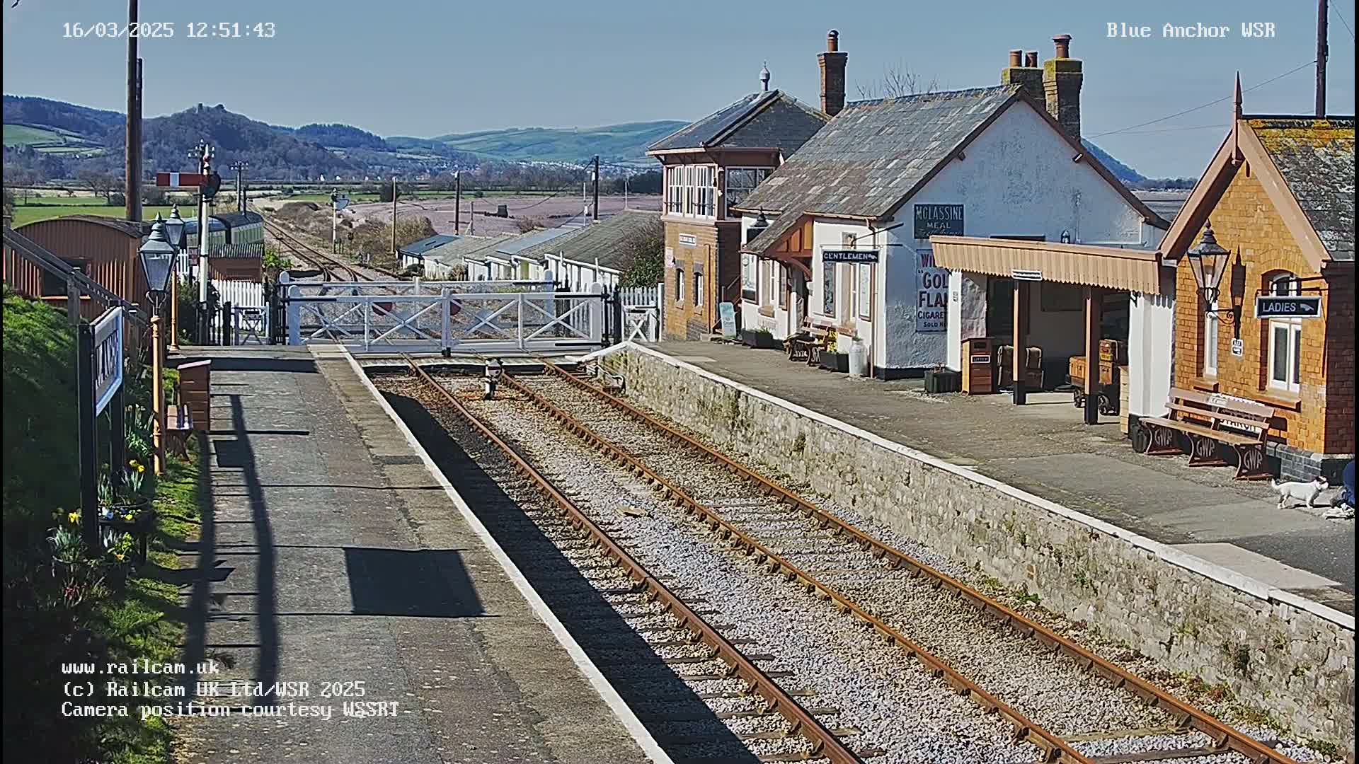 A sunny view of a train station with multiple tracks, a signal box, and station buildings, set against a backdrop of hills.