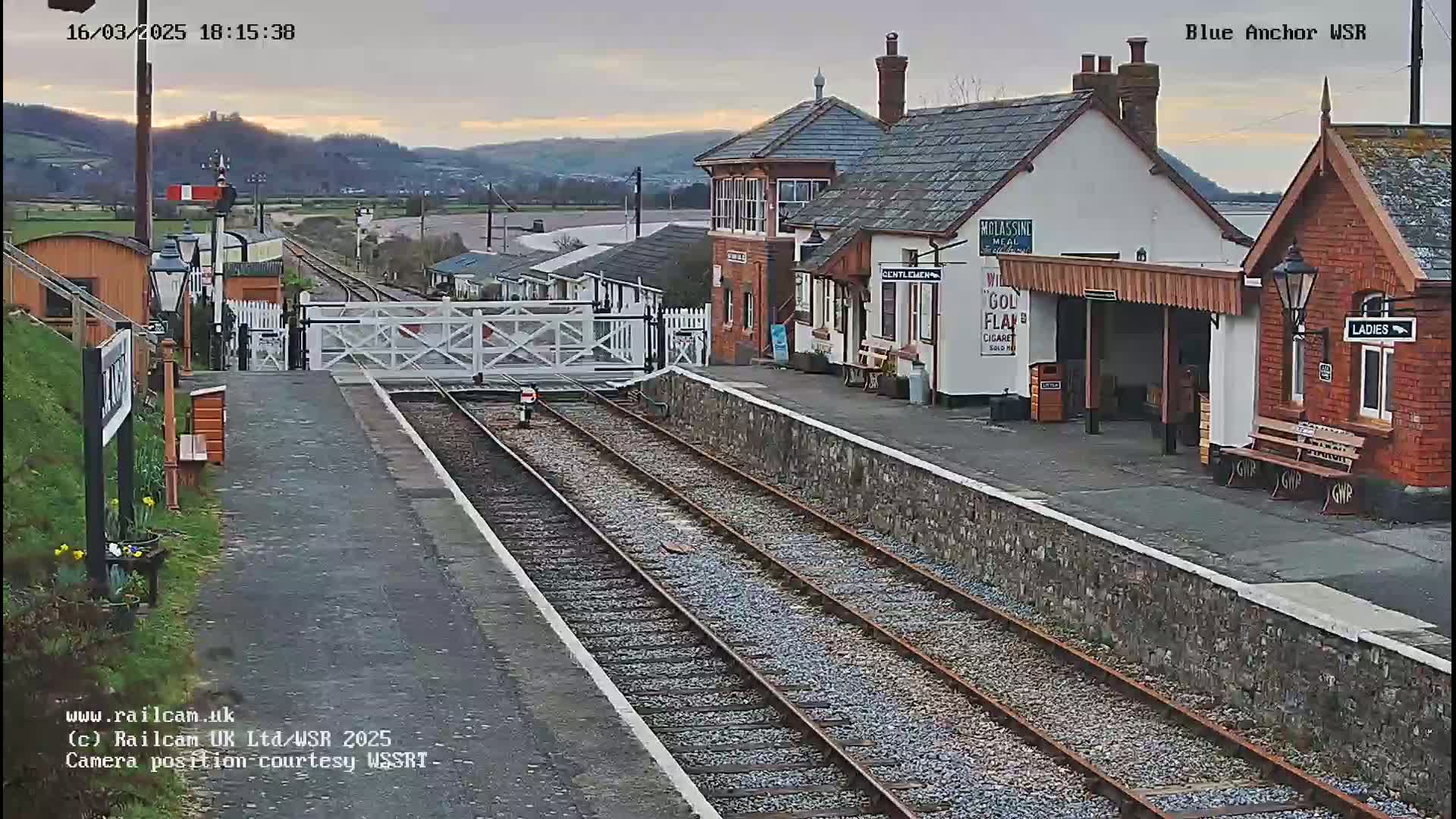 A railway station with two platforms and several buildings, under a partly cloudy evening sky, is viewed from the end of one of the platforms.