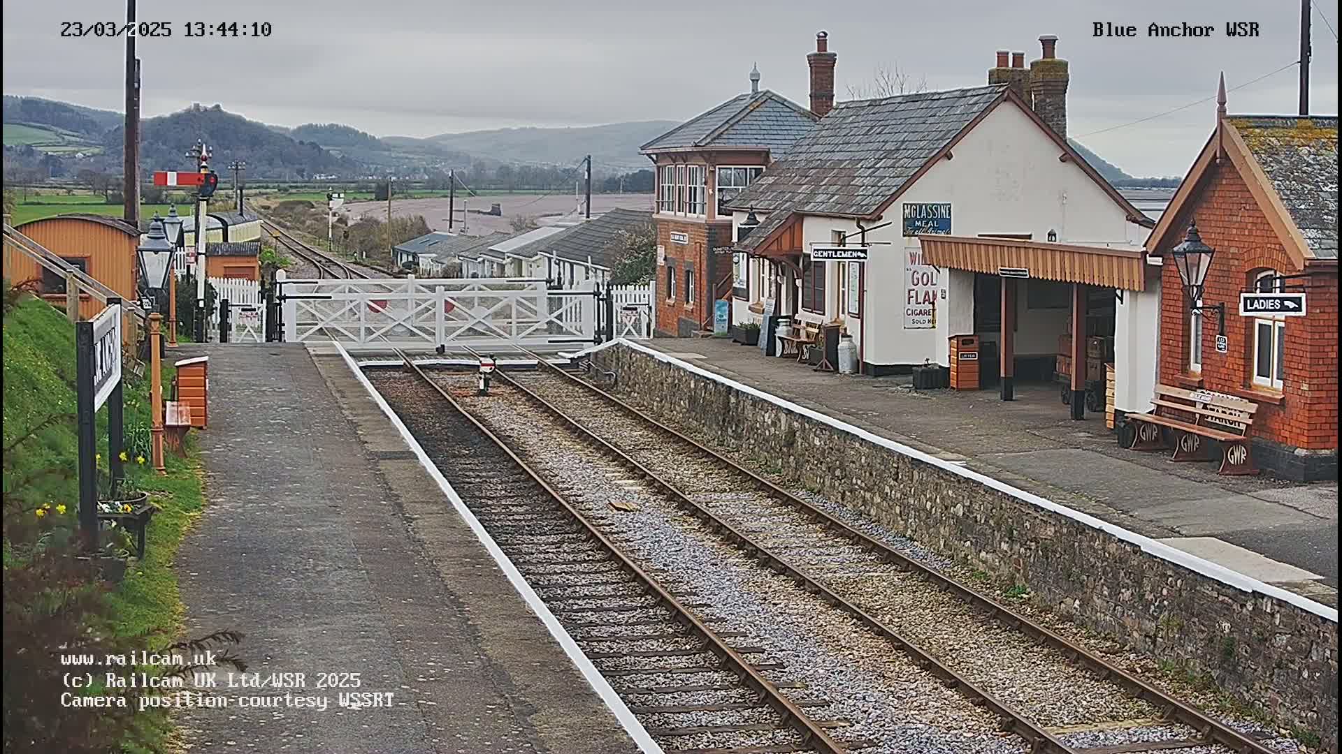 A railway station with a gray overcast sky, featuring two parallel train tracks leading to a platform with small buildings and benches.