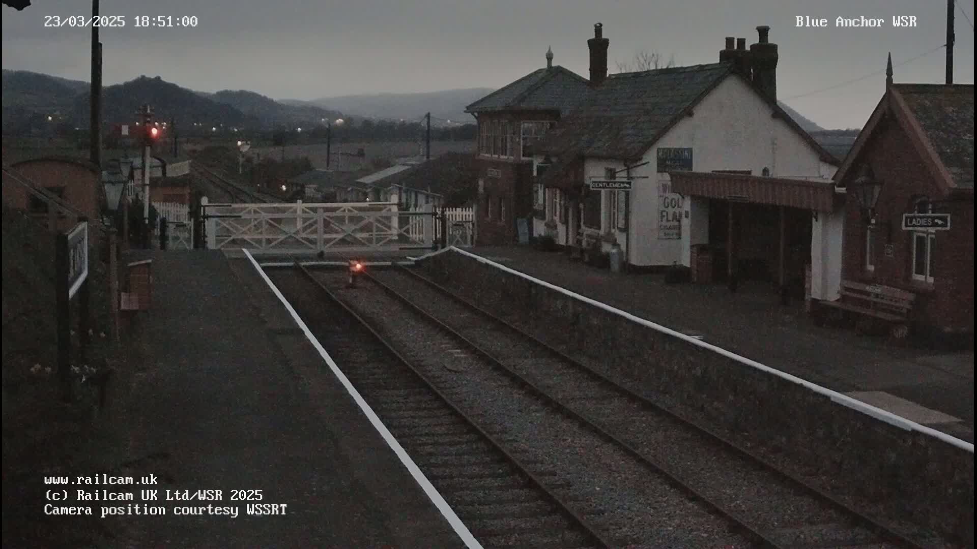 A train station with a low-lit platform, railroad tracks, and buildings under a dusky sky.