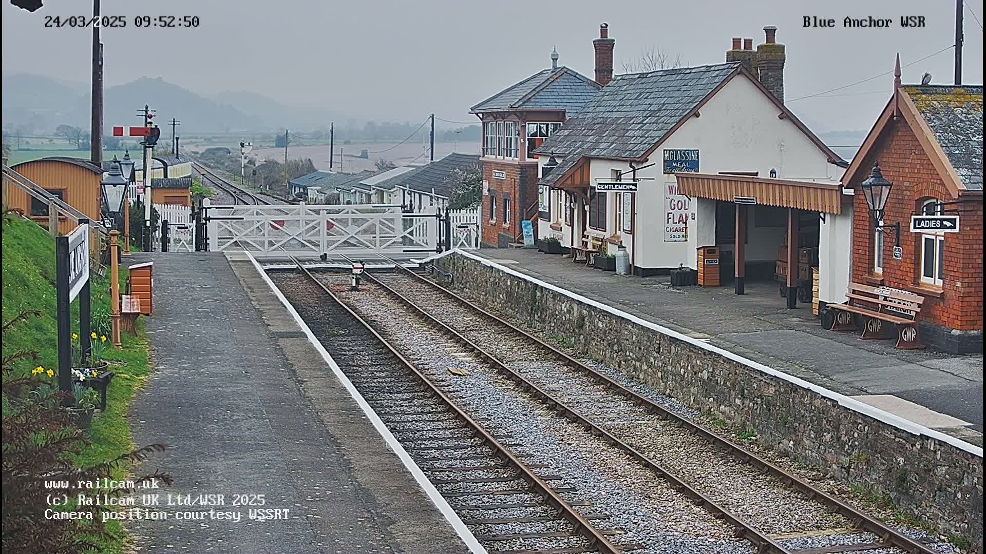 A train station with two passenger waiting areas and a signal box is viewed from the tracks on an overcast day.