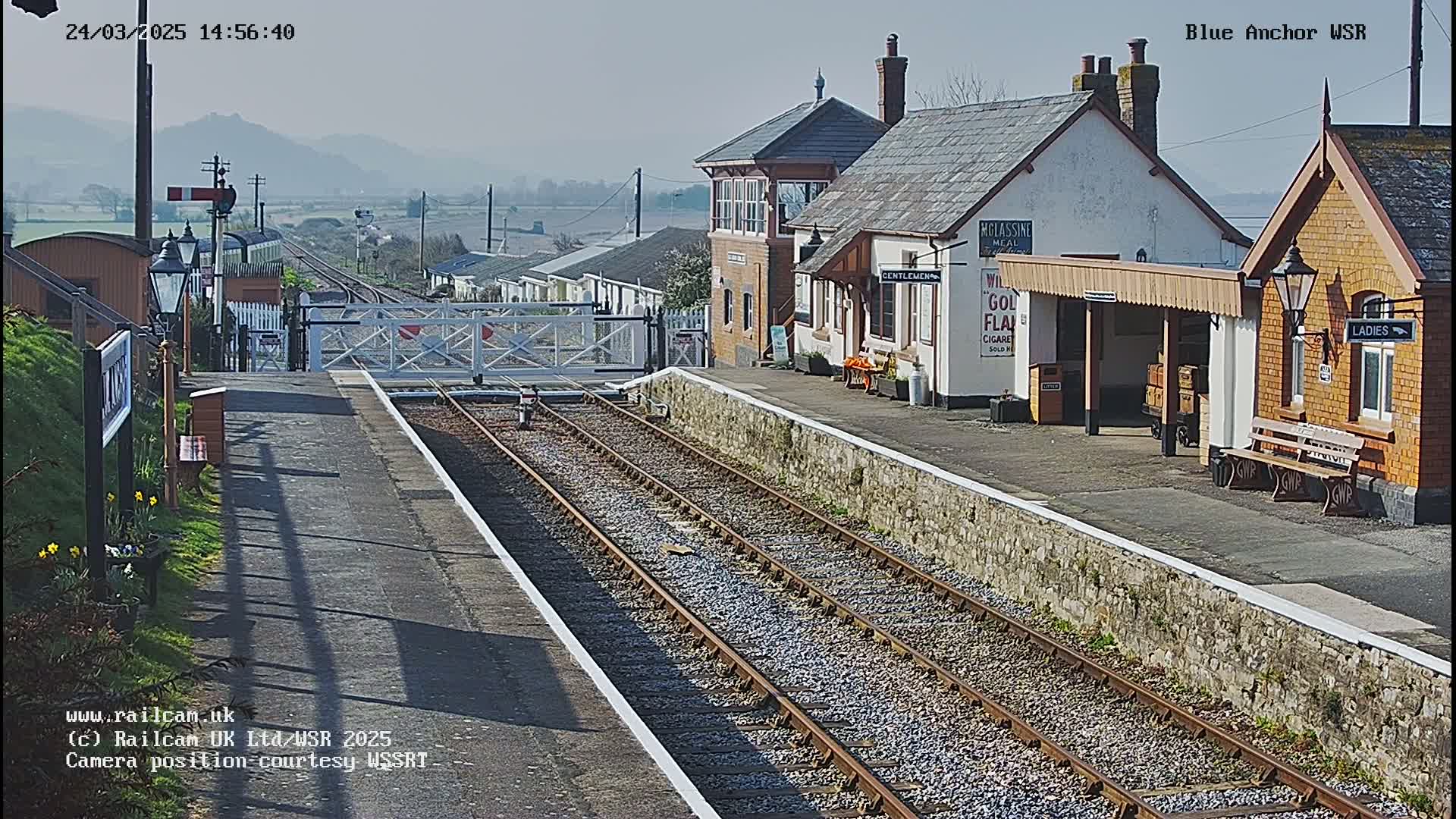 A sunny day at a train station with two parallel sets of tracks, a signal box, and passenger waiting shelters.