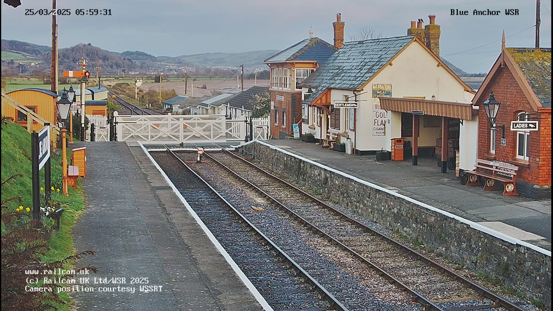 A train station platform with two sets of train tracks, a signal gate, and several small buildings under a cloudy sky.