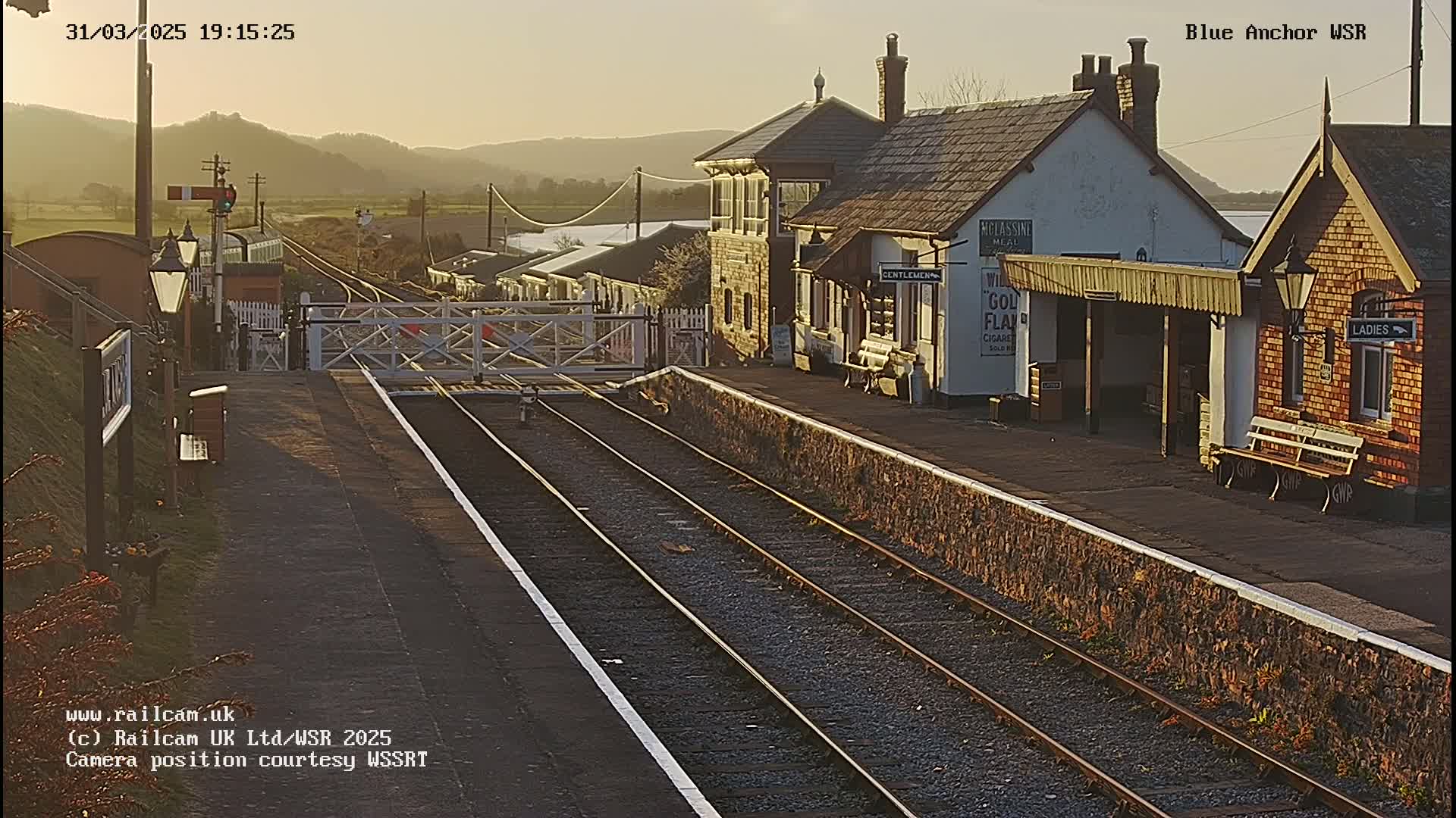 A train station with two platforms and several buildings, bathed in the warm light of the setting sun, sits next to train tracks.