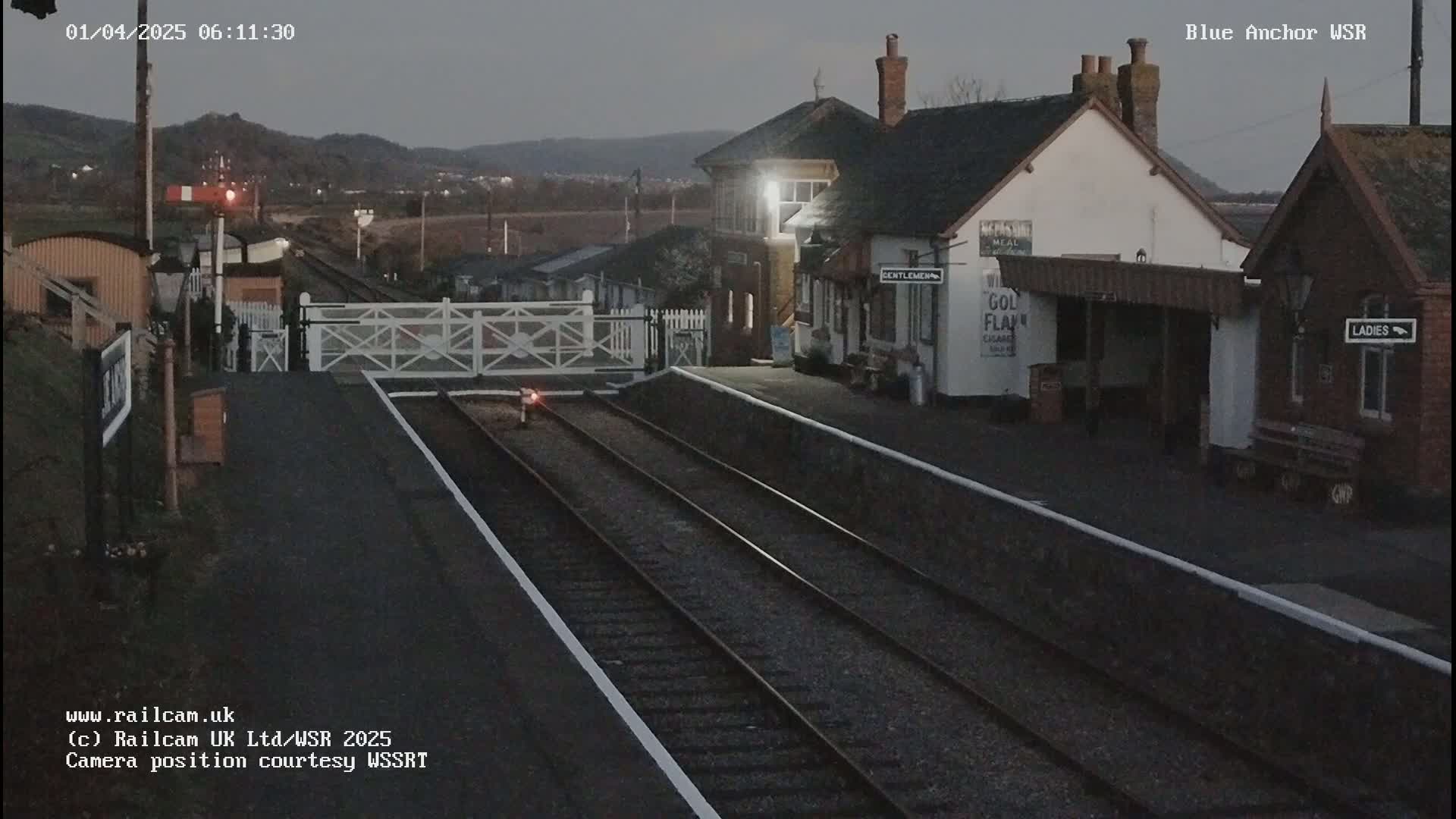 A dimly lit train station at twilight, with a level crossing gate down, and train tracks extending into the distance.