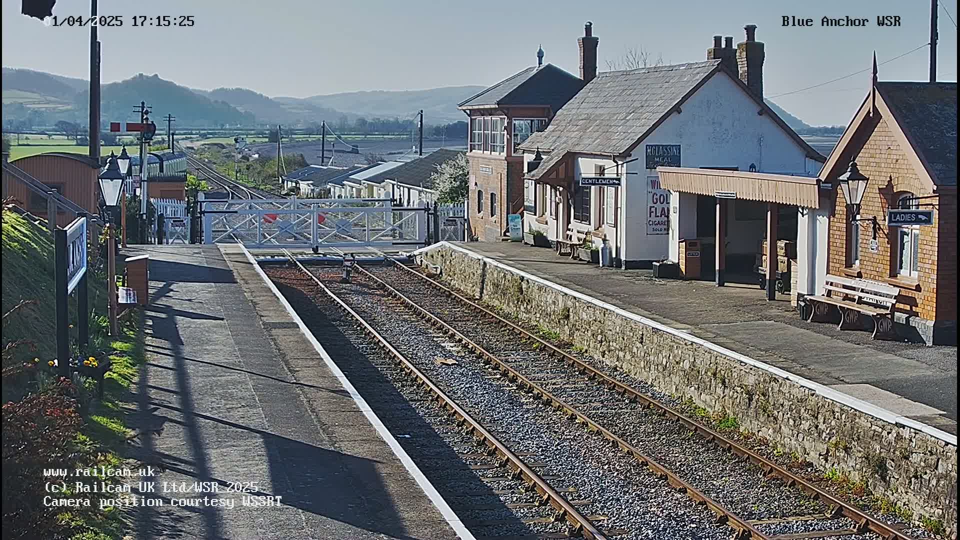 A sunny day at a train station with two parallel sets of tracks, a signal box, and passenger shelters.