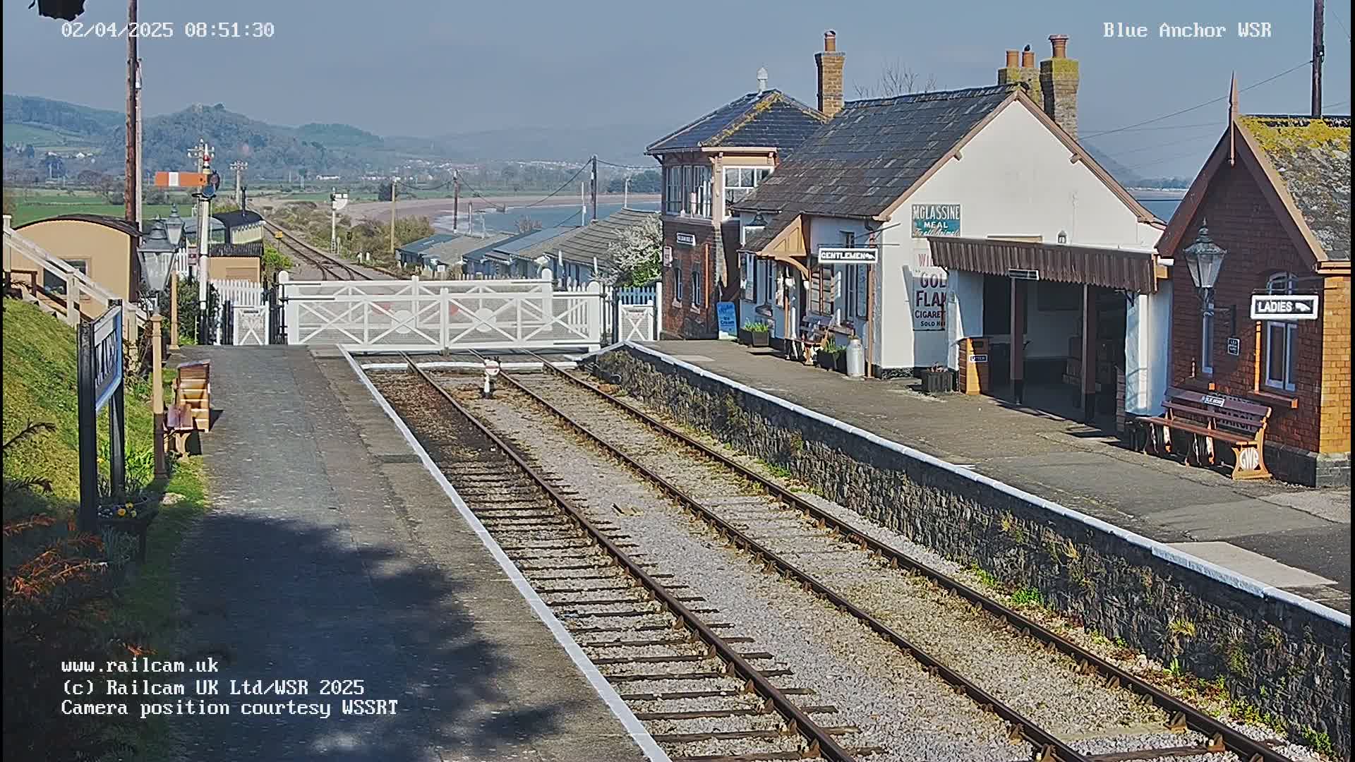 A sunny view of a railway station with two platforms, a waiting area, and a level crossing gate, situated near a body of water and hills under a clear sky.