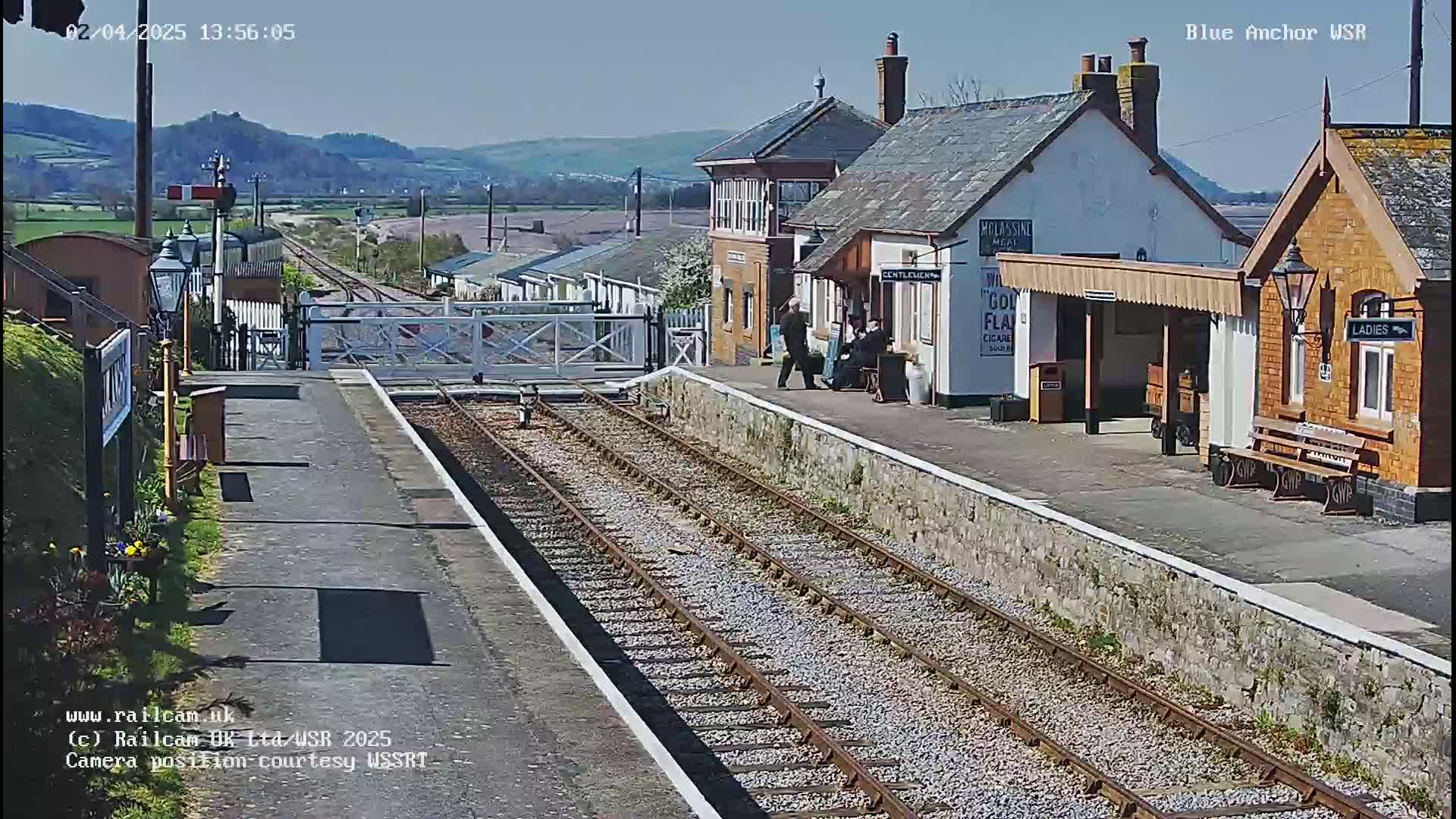 A sunny day at a train station with multiple buildings, a level crossing, and train tracks extending into the distance, with hills visible in the background.