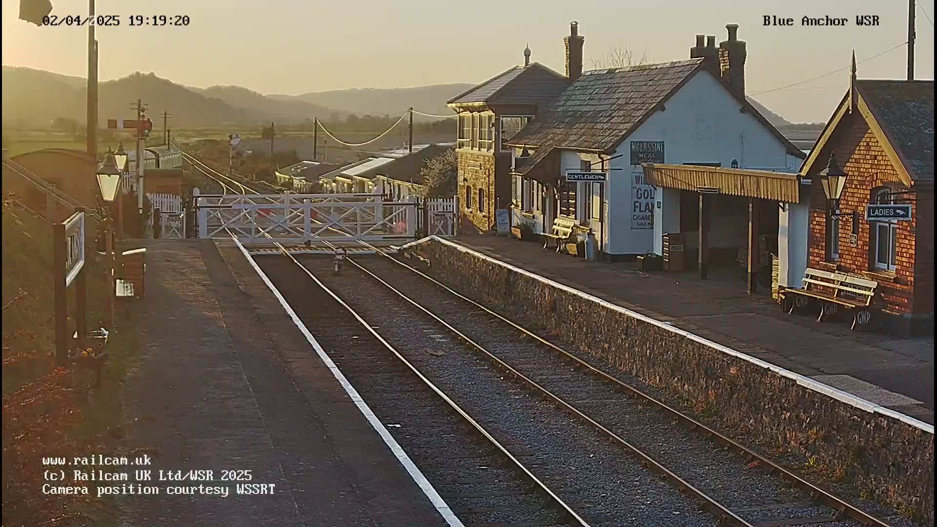 A train station with two platforms and several buildings is bathed in the golden light of sunset.