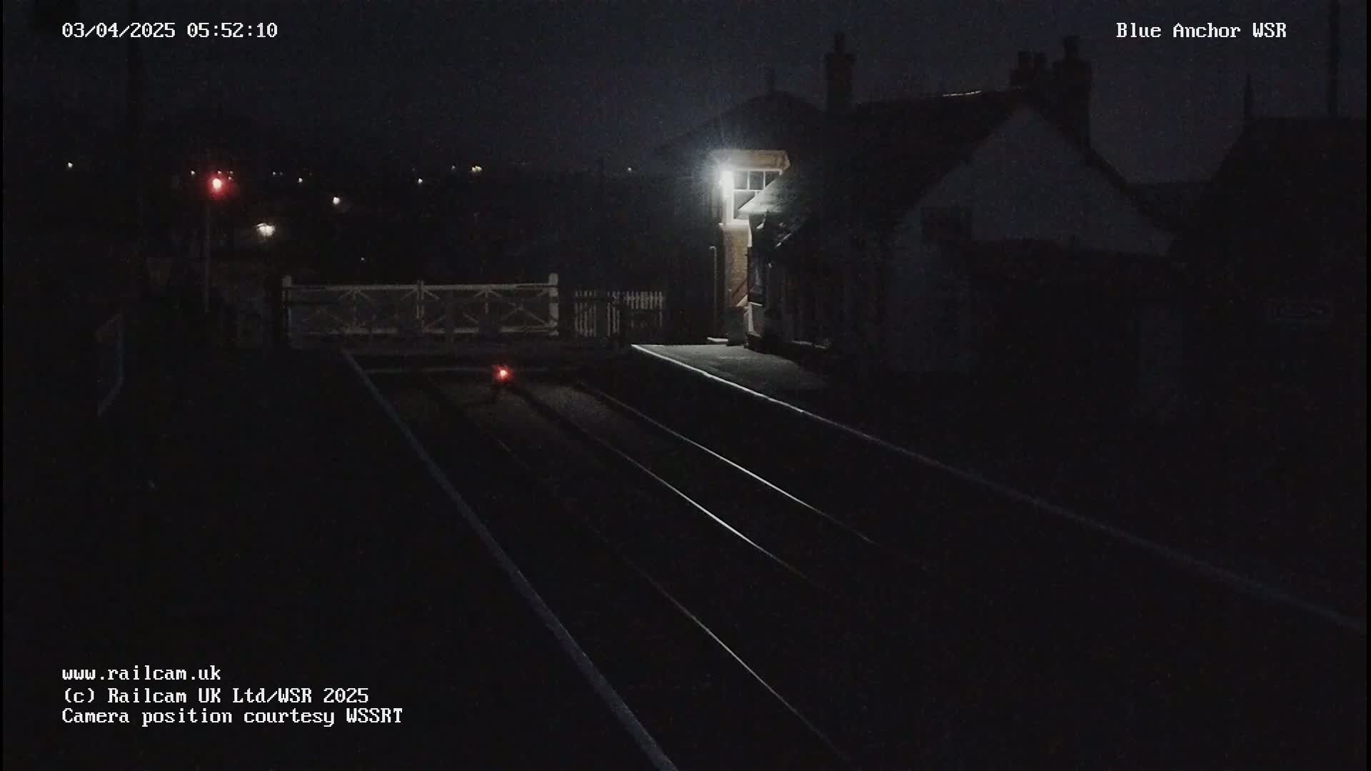 A dark, nighttime view of train tracks leading to a dimly lit train station building under a clear sky.