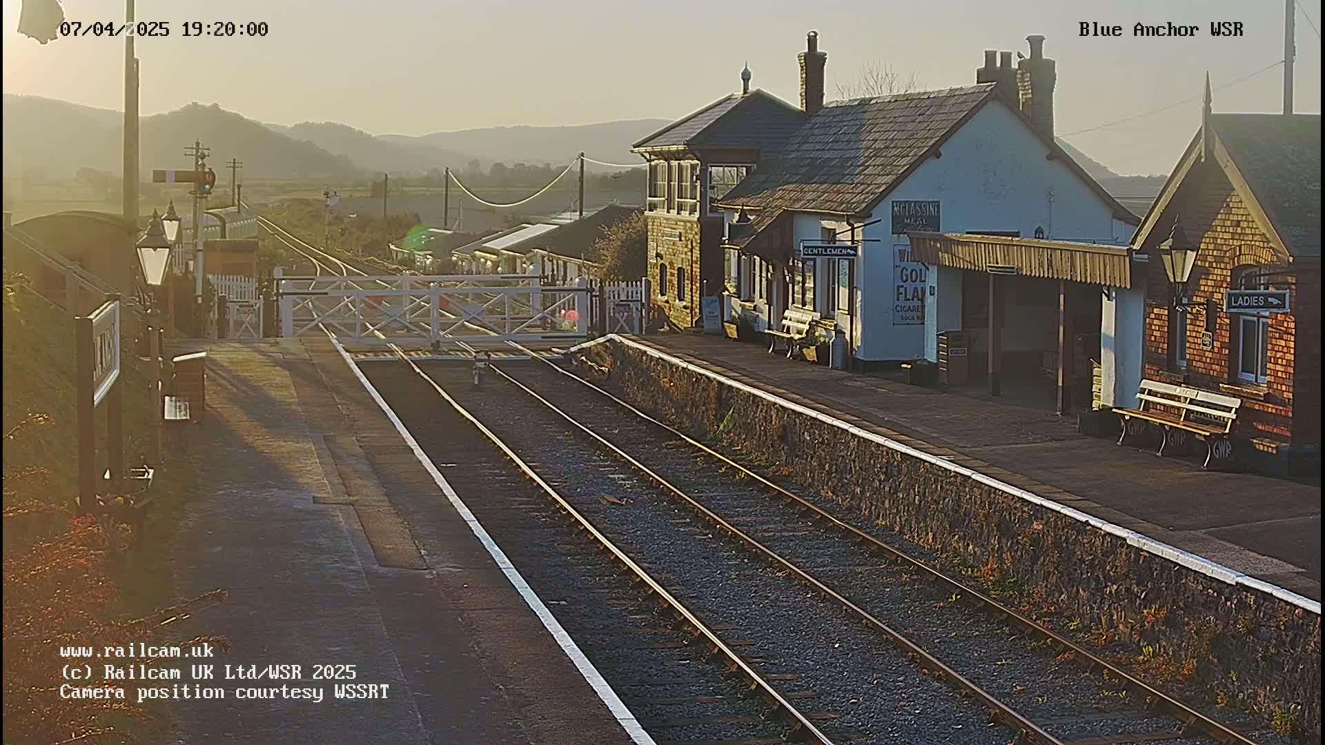 A train station platform with two sets of tracks and station buildings is seen at sunset against a backdrop of hills.