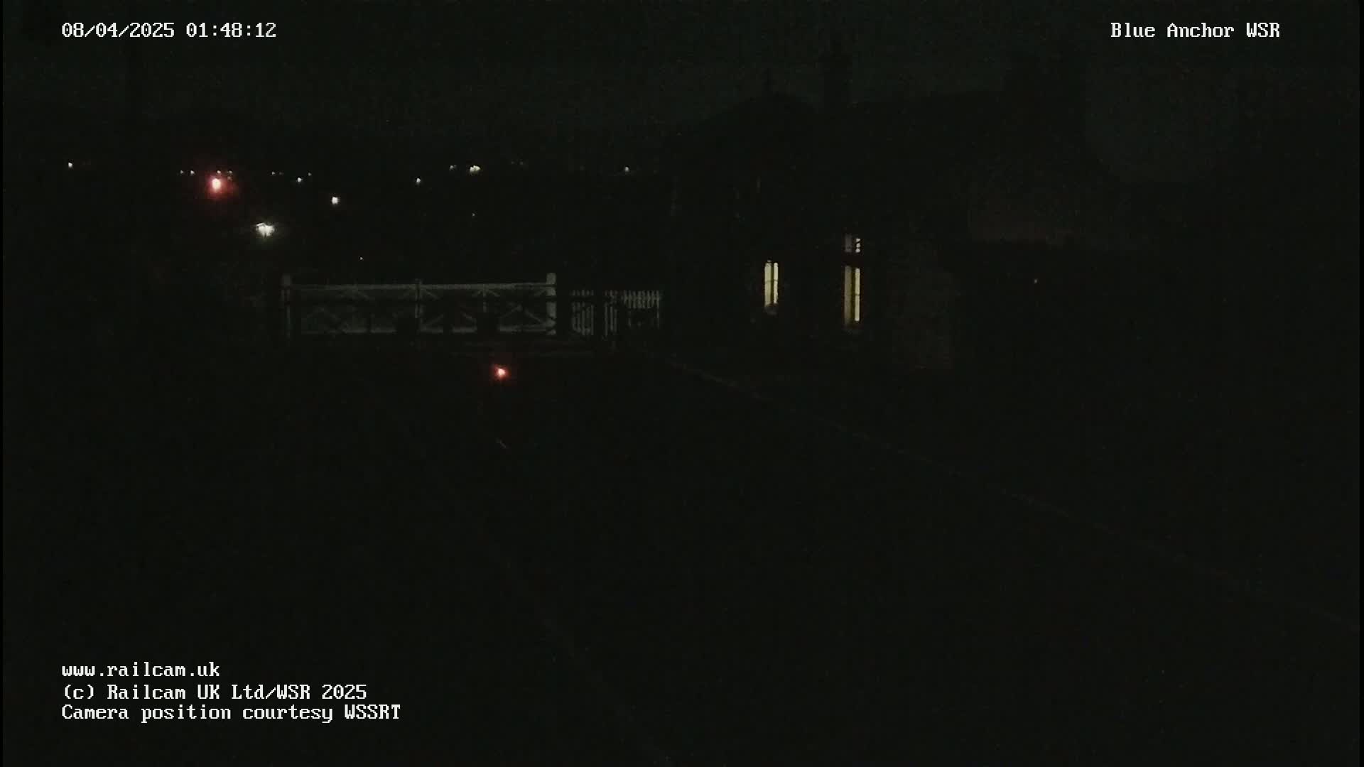 A dark, nighttime scene shows a dimly lit building and a fence near a railway track under a clear, dark sky.
