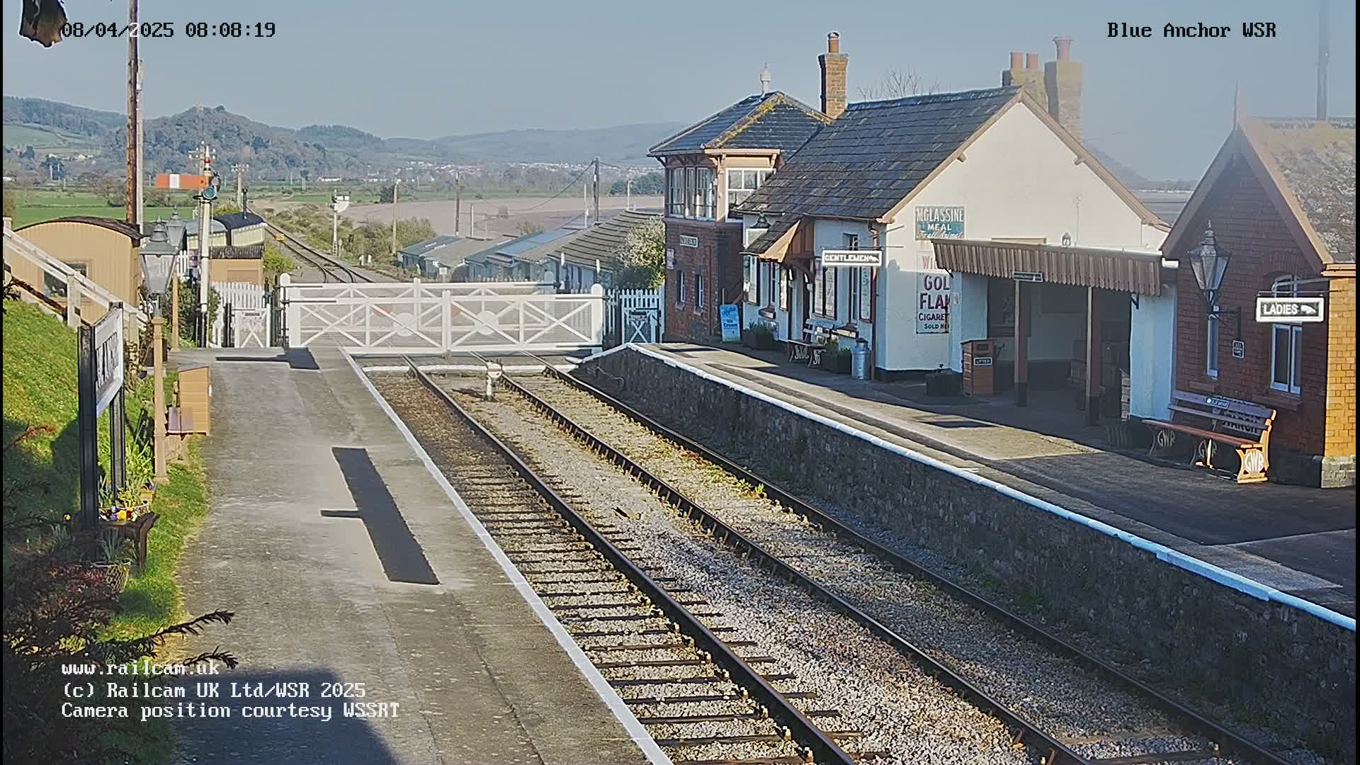 A sunny view of a train station with two sets of tracks, a signal gate, and several buildings, including a small station house and waiting shelters.