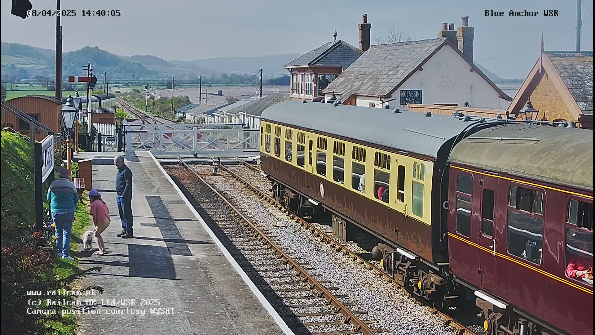 A maroon and cream passenger train is at a station on a sunny day, with several people standing nearby, one of whom is accompanied by a small dog.