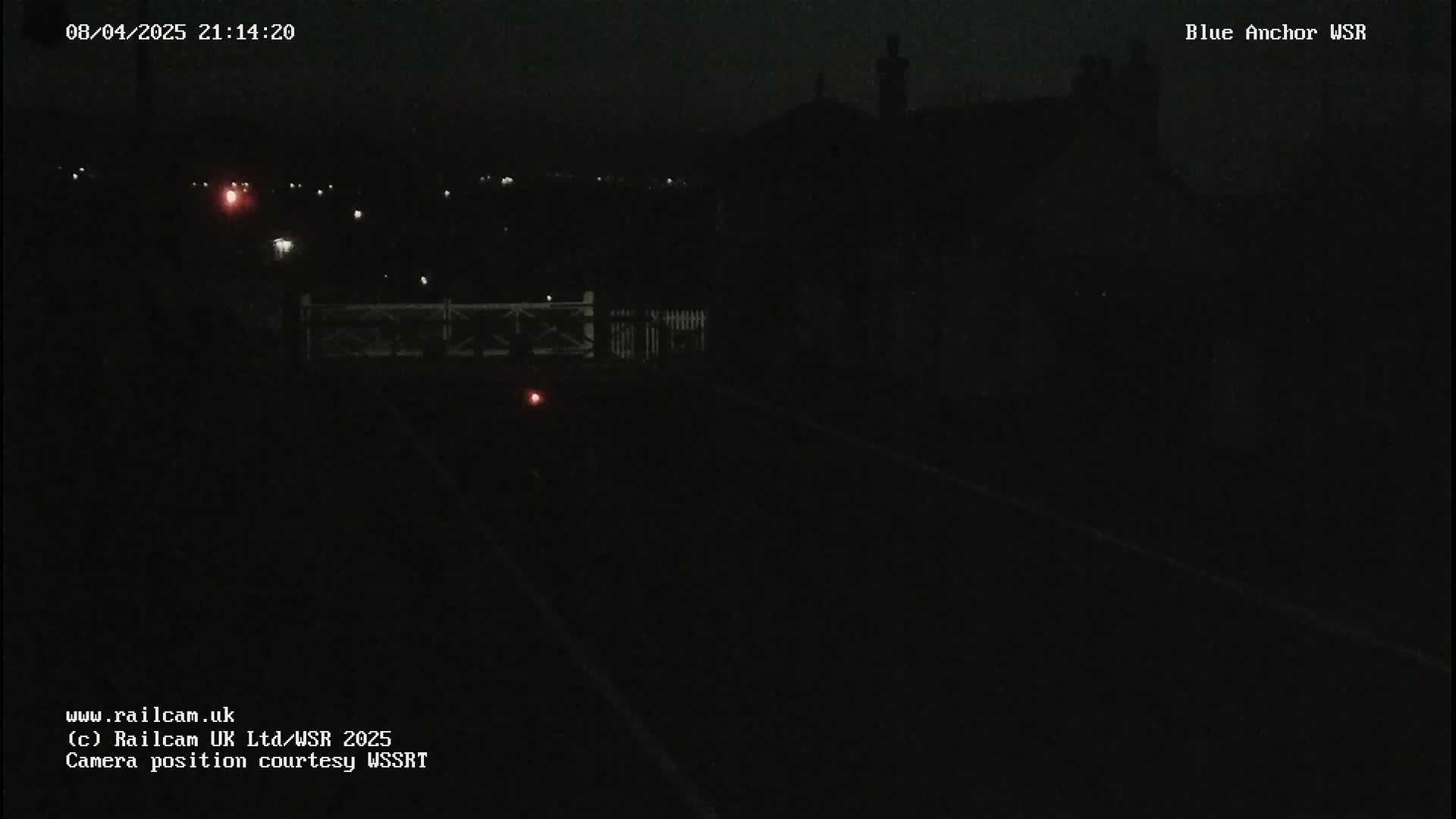 A dark nighttime scene shows a railroad track with a dimly lit building and a distant, faint light source in the background.