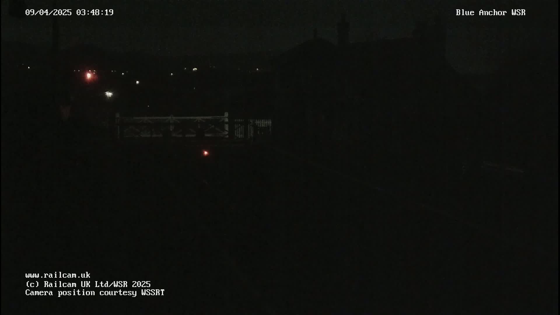 A dark nighttime scene shows a few faint lights in the distance, a dark structure in the middle ground, and a dark fence or railing in the foreground.