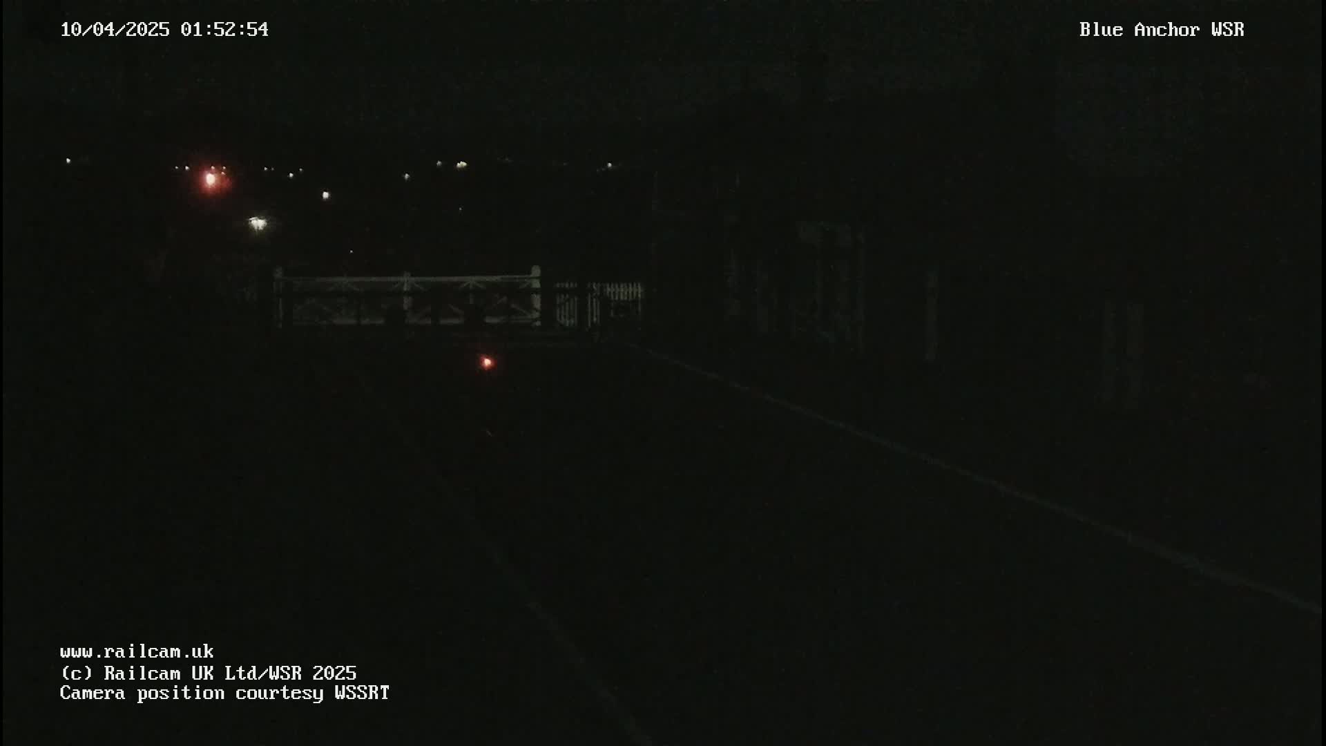 A dark, nighttime view of a roadway leading to a gated crossing, with faint lights visible in the distance.